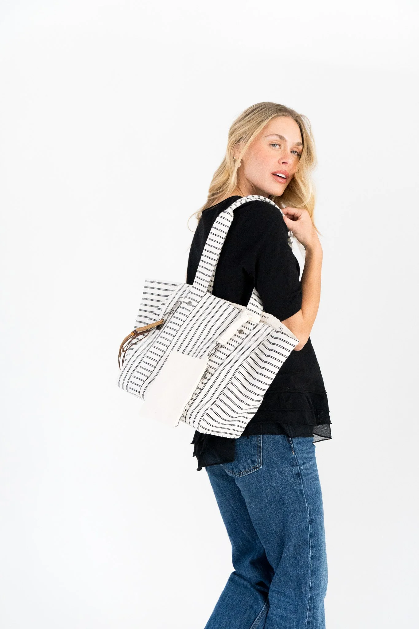 A woman with blonde hair wearing a black top and blue jeans, holding a white striped tote bag, posing against a plain white background.