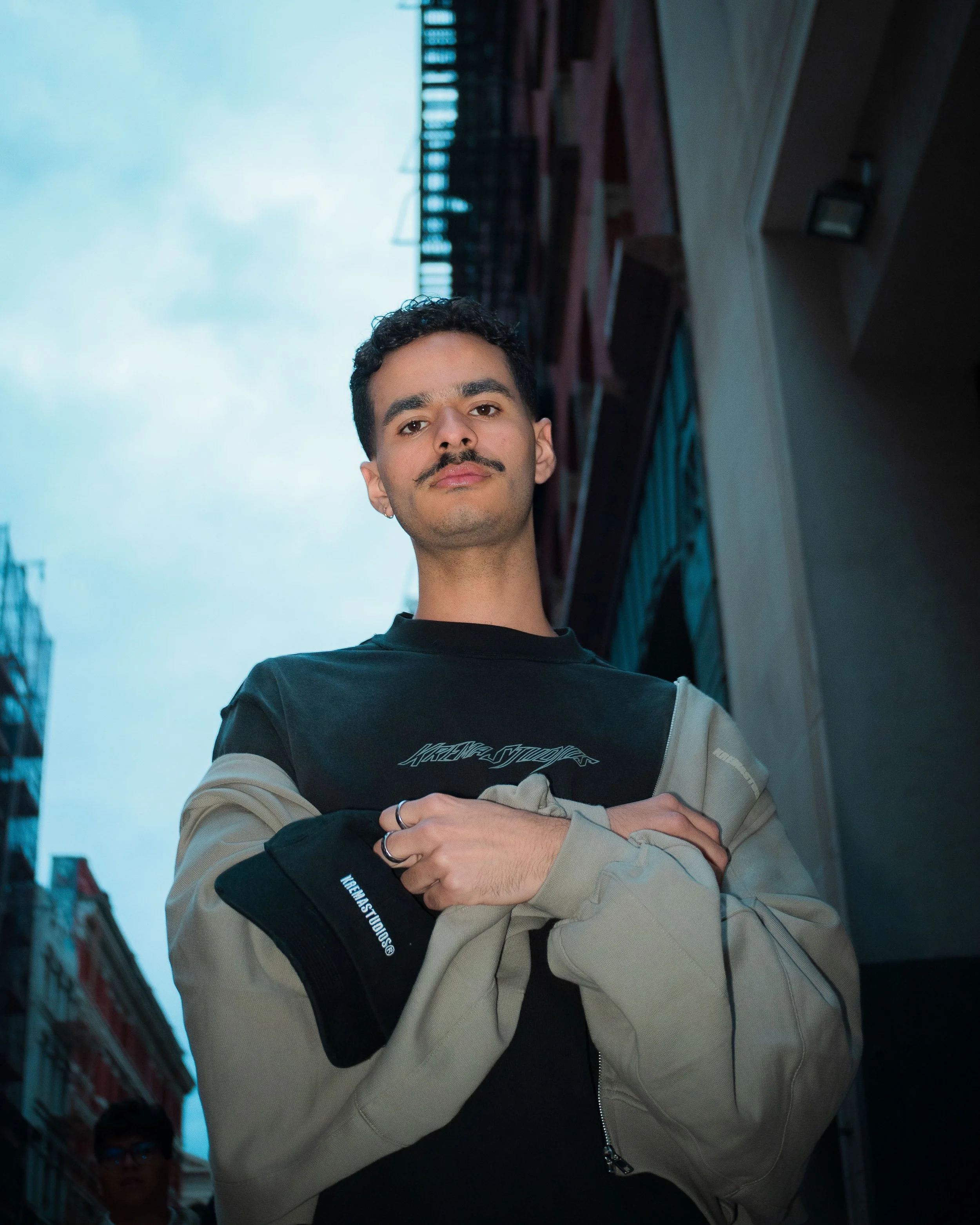Young man with curly hair and mustache standing on city street with buildings and cloudy sky behind him, wearing a black t-shirt and beige jacket, holding a black cap.