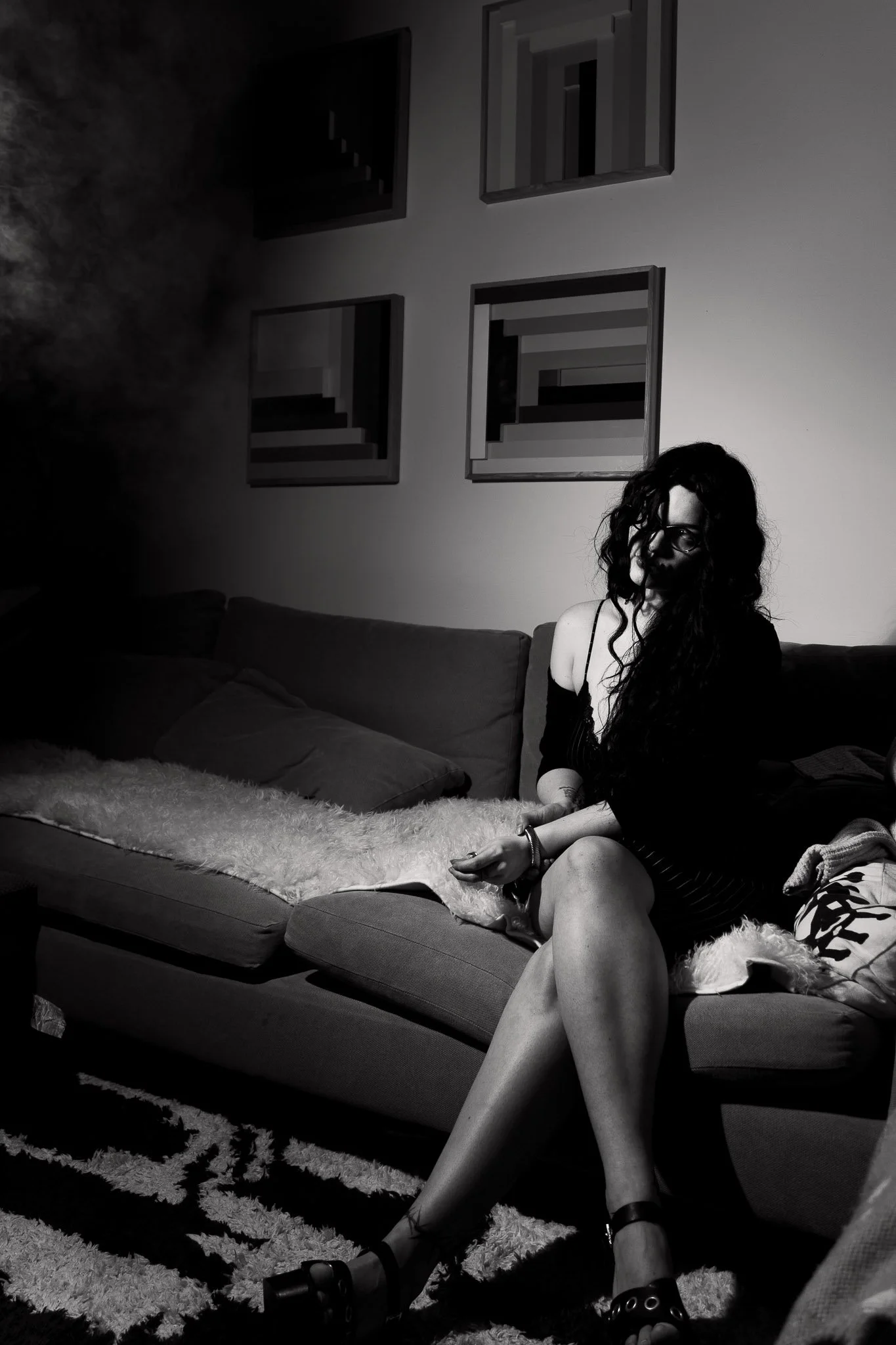 Black and white photo of a woman sitting on a sofa in a modern living room. The woman has long, curly hair, glasses, and is wearing a dark dress. She is resting her arm on her knee and looking towards the camera. The sofa has several pillows and a fl