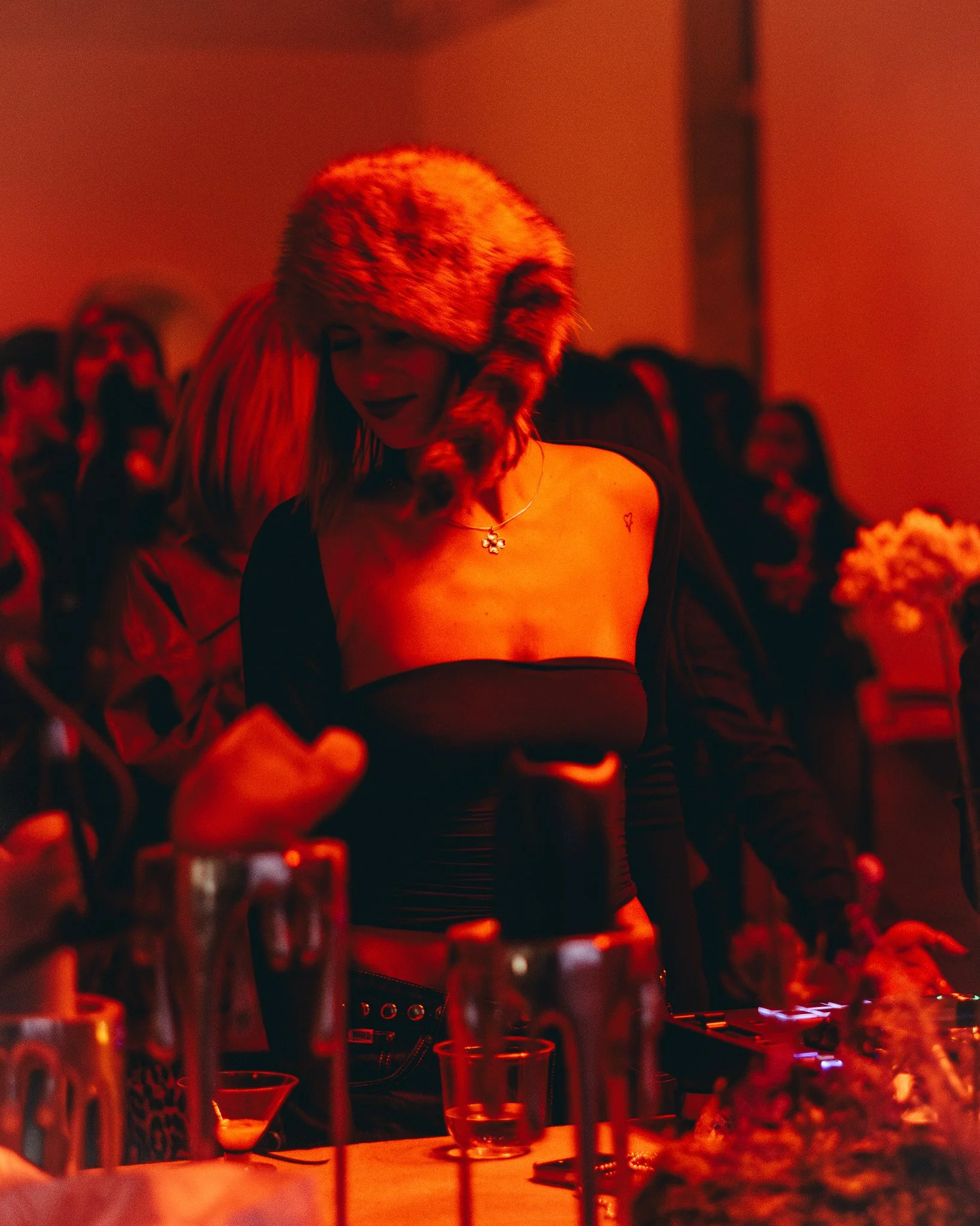 A woman with a furry hat with ear flaps, wearing a black top with a necklace, appears to be DJing at a party or nightclub with red lighting. Several people in the background are seen, and there are drinks and glasses on the table in front of her.