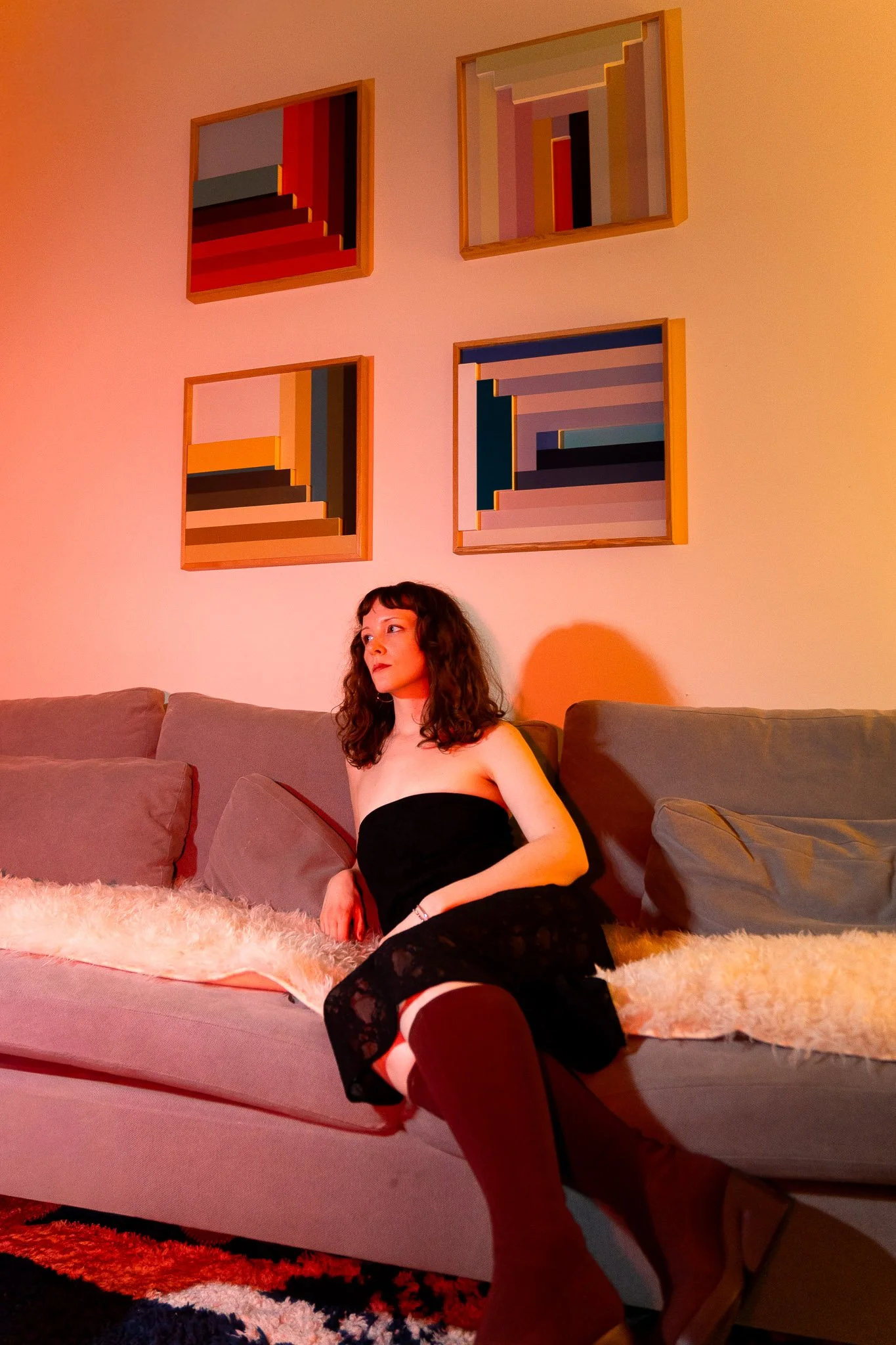 A woman with curly dark hair, wearing black lace lingerie, black thigh-high stockings with lace tops, and white and red garter straps, sitting on a couch with a beige blanket. Behind her on a beige wall are four colorful, abstract, geometric painting