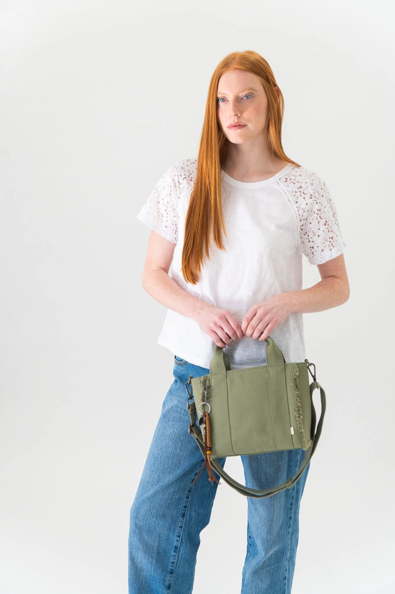 A woman with long red hair wearing a white lace sleeve top and blue jeans, holding a green handbag with decorative charms, standing against a plain white background.