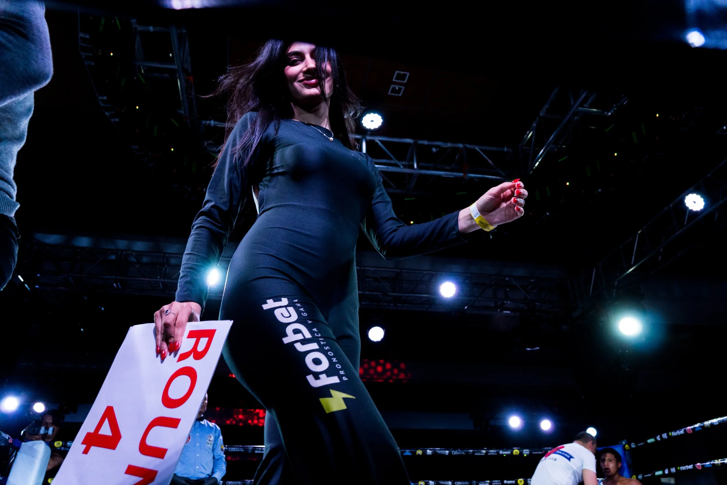 A woman in a dark athletic outfit with yellow accents, dark hair, and light skin is smiling and holding a white paper bag, standing in a boxing ring with bright lights overhead.