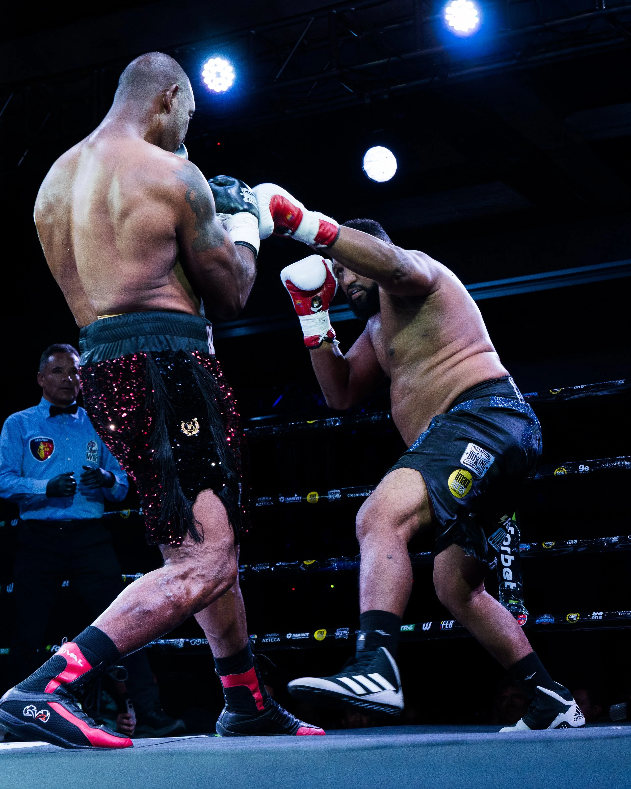 Two male boxers in a boxing ring during a match, with one delivering a punch to the other, and a referee in the background observing.