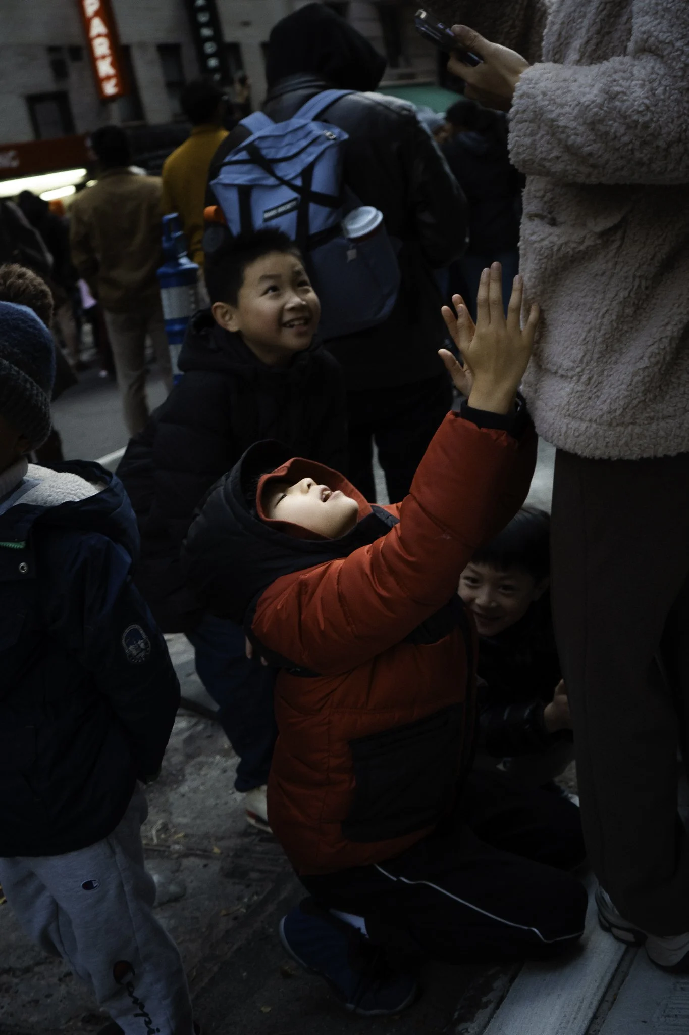 A group of children and adults standing on a city street, with some wearing backpacks and jackets. One child in a red jacket is kneeling on the ground touching a man’s leg, looking up.