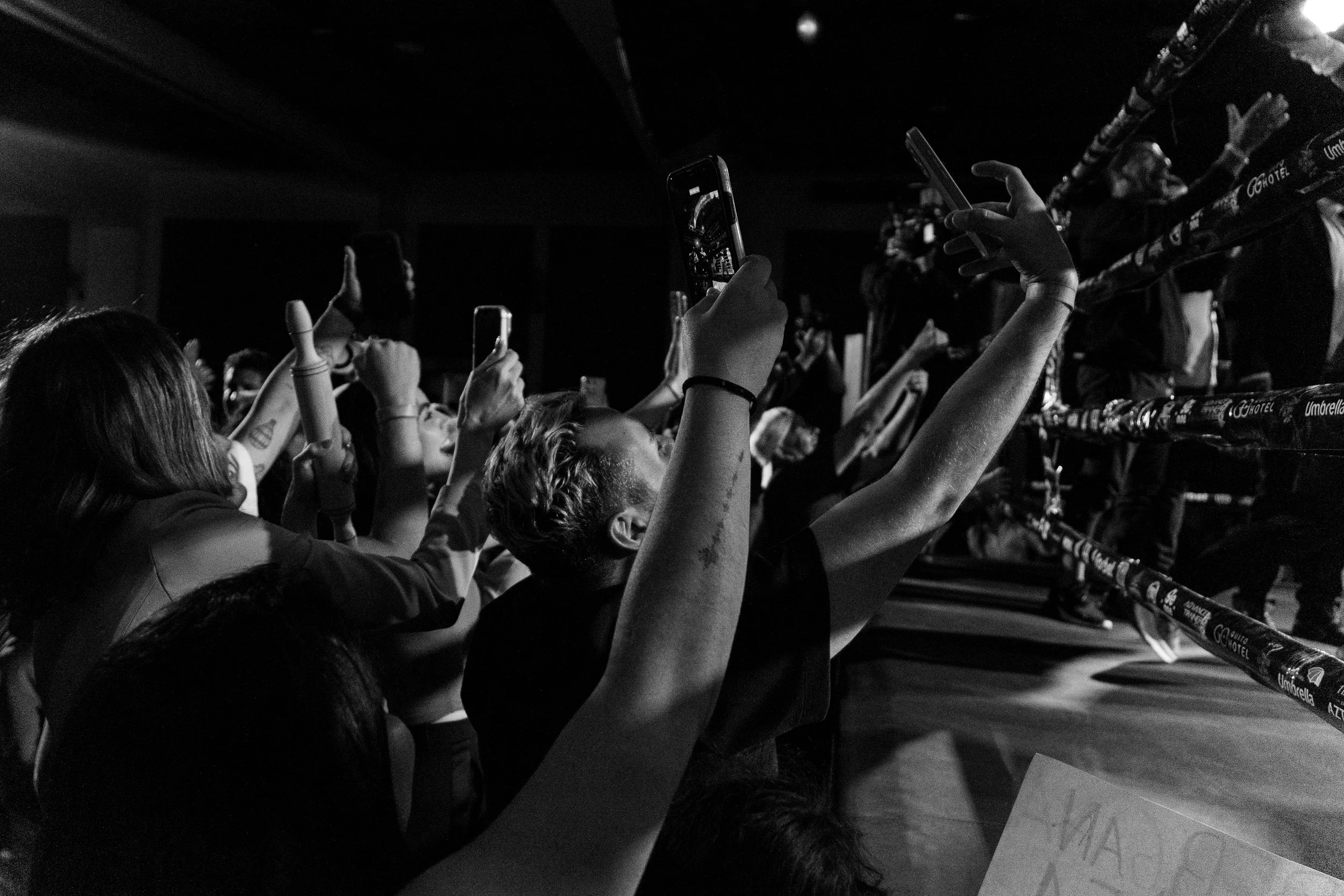 A crowd of people at an event, raising their hands and taking photos with smartphones, with a row of umbrellas visible in the background.