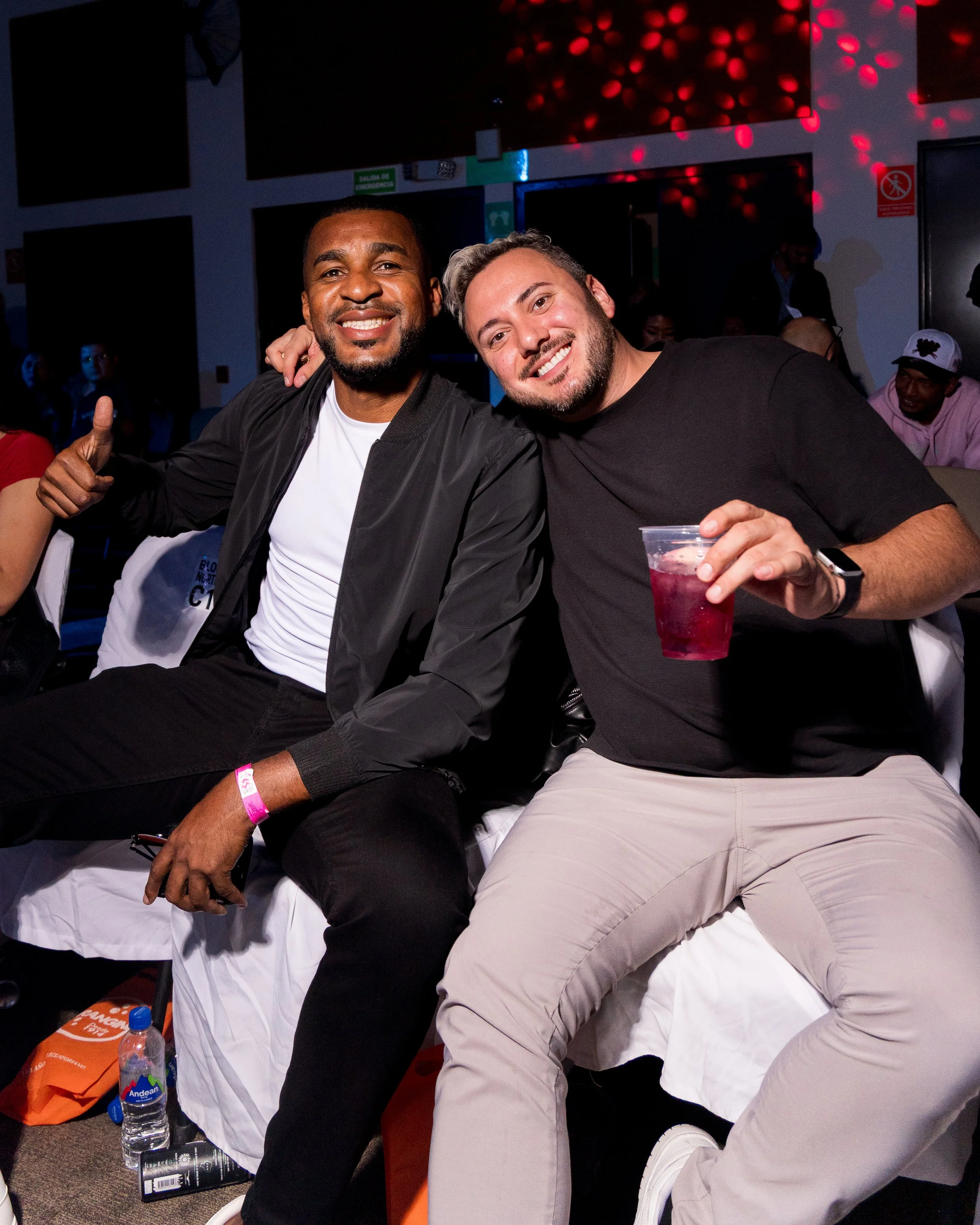 Two men sitting closely, smiling at the camera. One is wearing a black jacket and white shirt, the other a black t-shirt and light-colored pants, holding a red drink in a clear cup. They are in a dark indoor setting with colorful lights and other peo