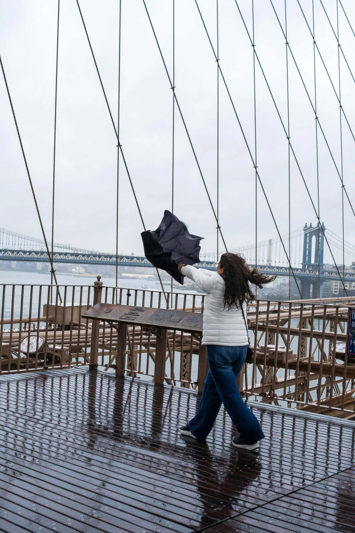 A woman in a white jacket and blue jeans on a wet wooden deck of a bridge, holding a black umbrella in the wind with the Manhattan Bridge in the background on a cloudy day.