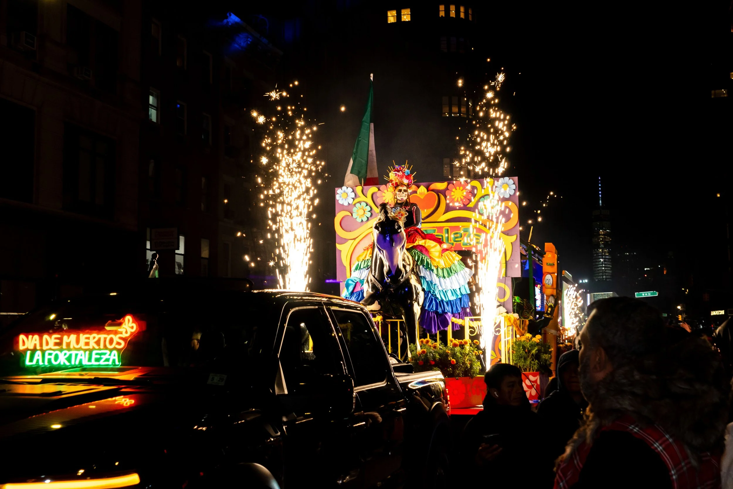 Nighttime parade with a float featuring a colorful figure dressed in traditional Mexican attire, wearing a skull face makeup, riding a black horse, with sparks flying from the sides, and a Mexican flag in the background. Neon sign reading "Día de Mue