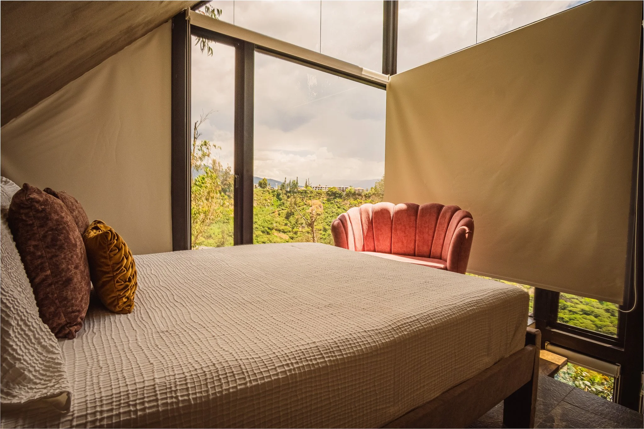 A cozy bedroom with a large bed covered in a textured beige bedspread, rose velvet pillows, and a mustard yellow pillow. There is a pink velvet armchair at the end of the bed. The room features large windows with a view of lush green trees and distan