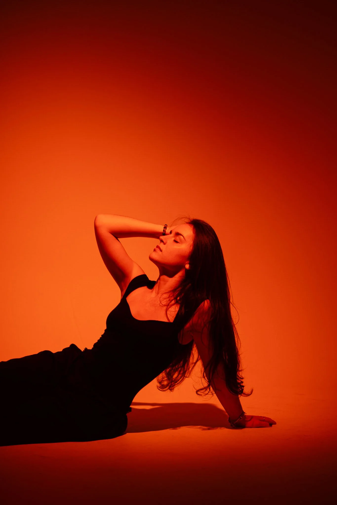 A woman with long dark hair sitting on the ground against a warm orange background, with her head tilted back, eyes closed, and one arm behind her head.