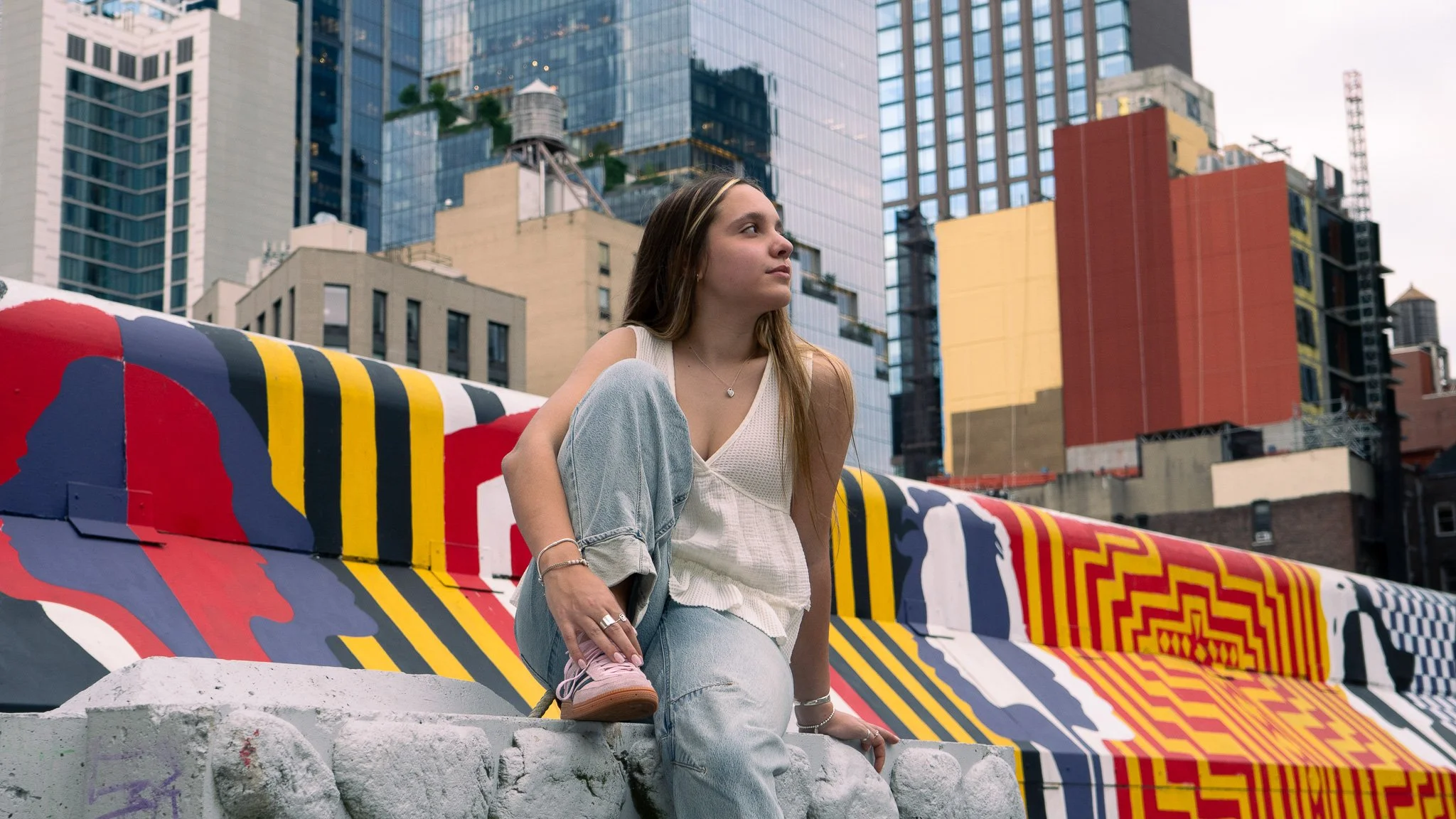 Young woman with long hair sitting on colorful painted concrete barrier in urban setting with tall glass skyscrapers behind her.