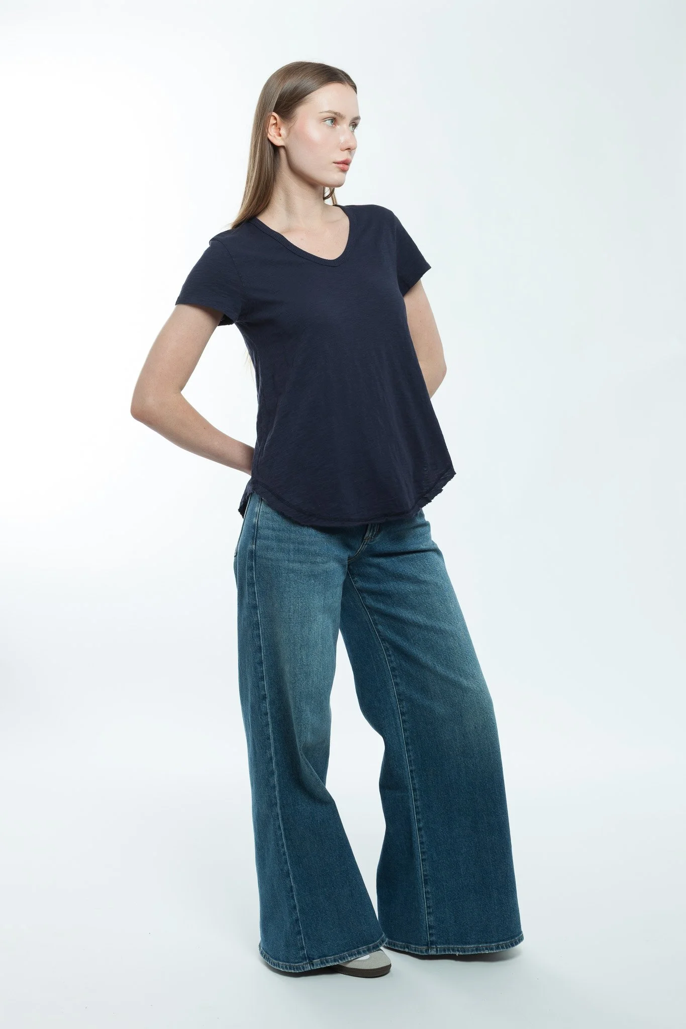 A woman with long brown hair wearing a navy T-shirt and wide-leg jeans, standing against a plain white background.