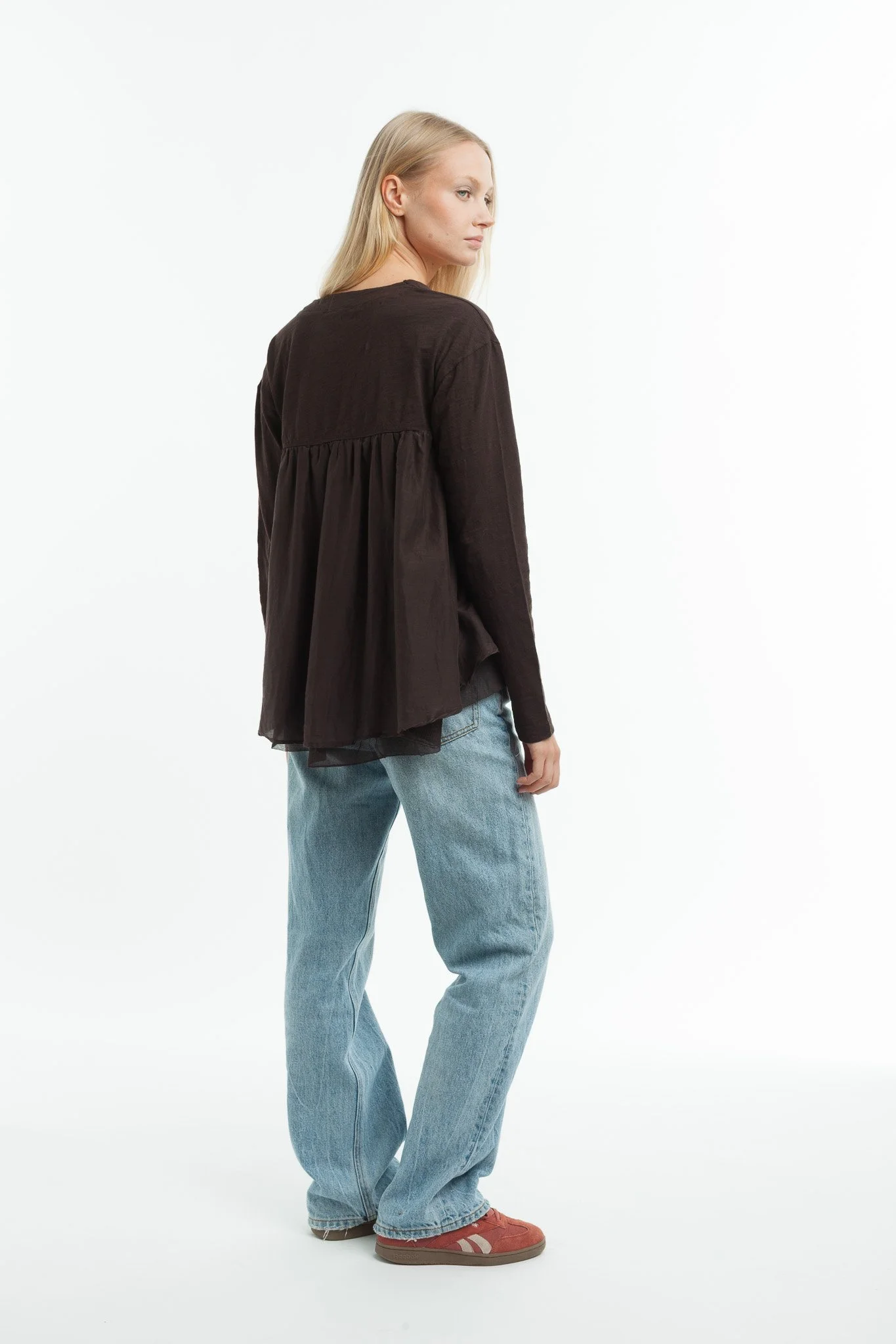 A woman with long blonde hair wearing a black loose top, light blue jeans, and red sneakers standing against a white background.