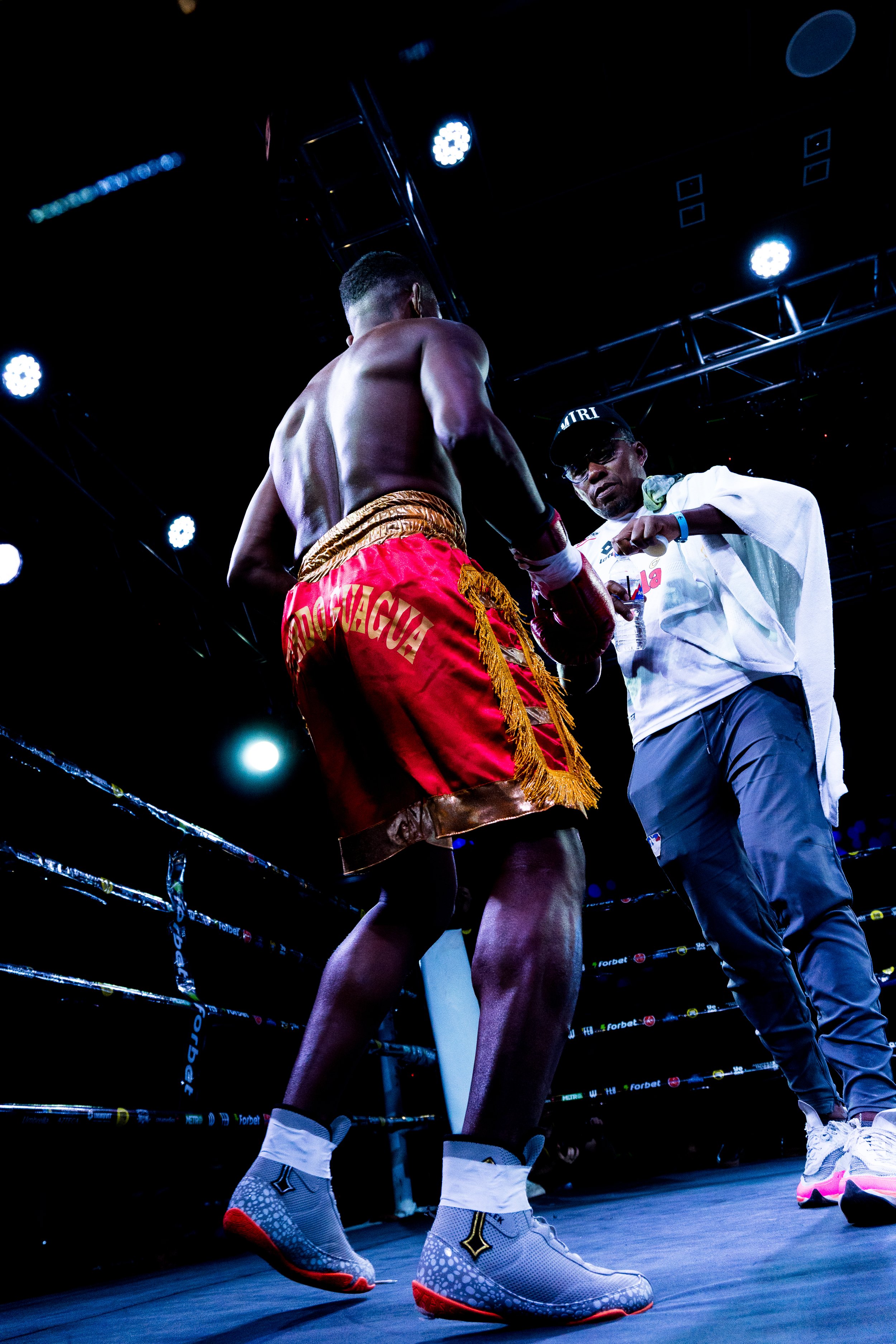 A boxer in red and gold trunks with the name "RODRIGUEZ" on the back standing in a boxing ring, receiving a thumbs-up from a man in a white shirt and cap, under bright lights with a dark background.