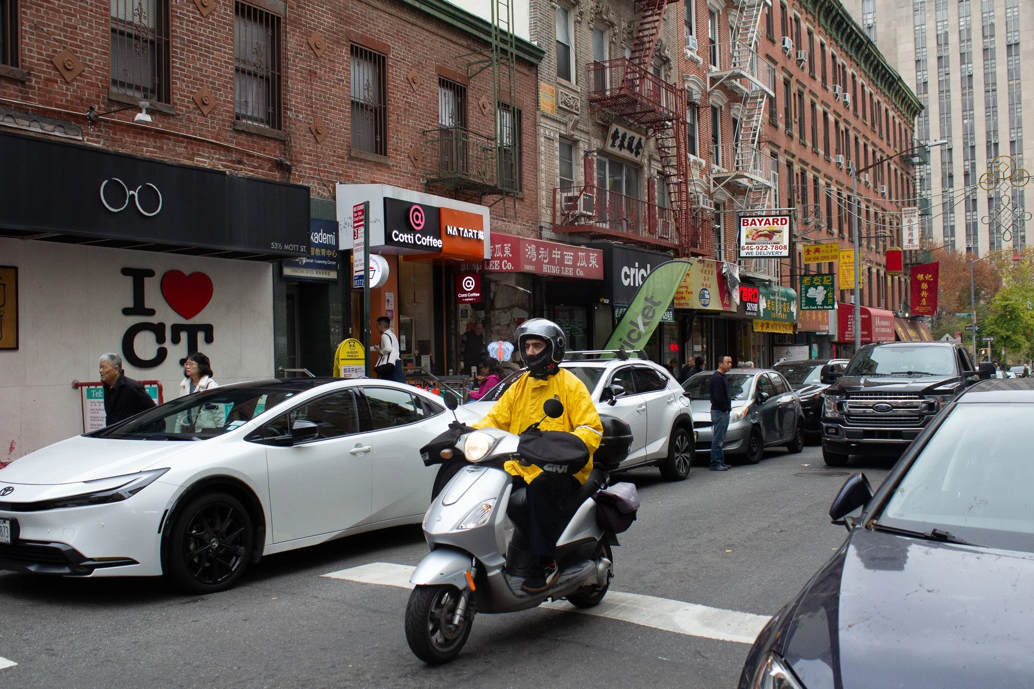 City street with parked cars and pedestrians, a delivery scooter with a rider in a yellow jacket, brick buildings with storefronts, signs in English and Chinese, trees in the background.
