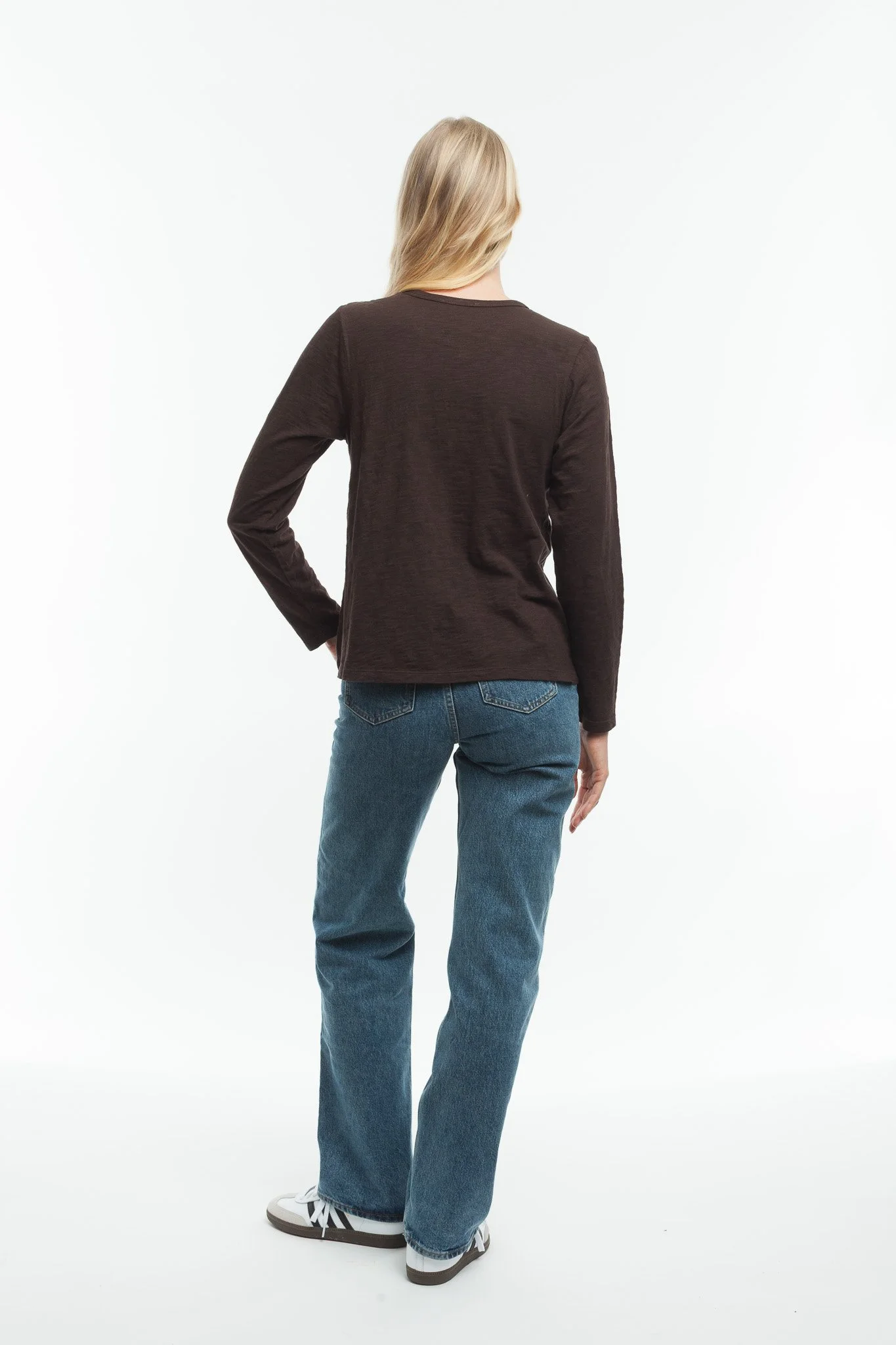 A woman standing with her back facing the camera, wearing a dark long-sleeve shirt, blue jeans, and sneakers, with a white background.