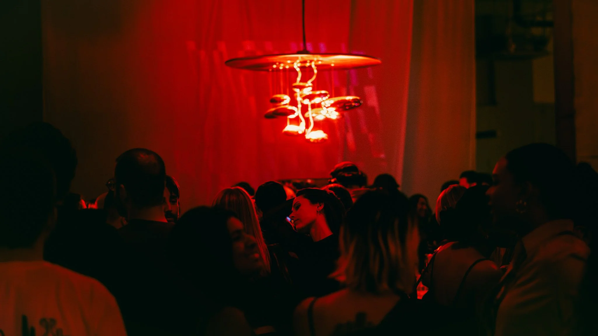 A dark room filled with people, illuminated by red lighting, with a modern, artistic hanging light fixture.