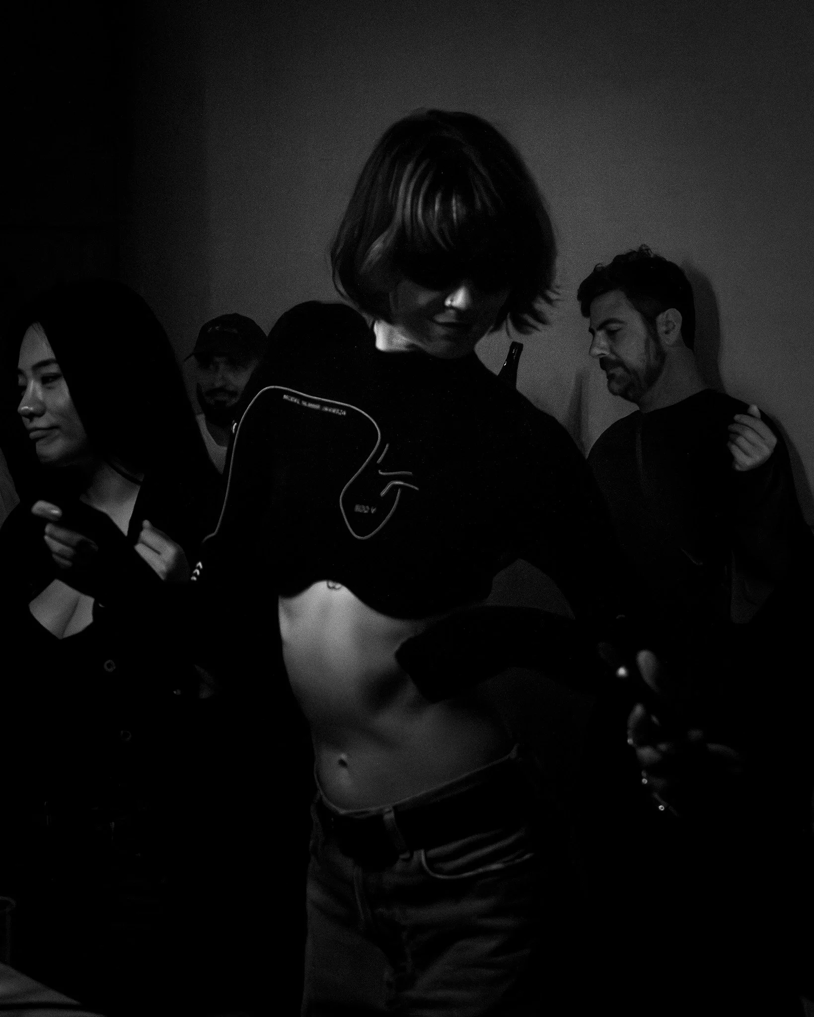 Black and white photo of multiple people, mostly men, with one woman visible, gathered together. One man in focus has a beard and short hair, and the woman has long dark hair, holding a phone. The setting appears to be indoors with a plain background