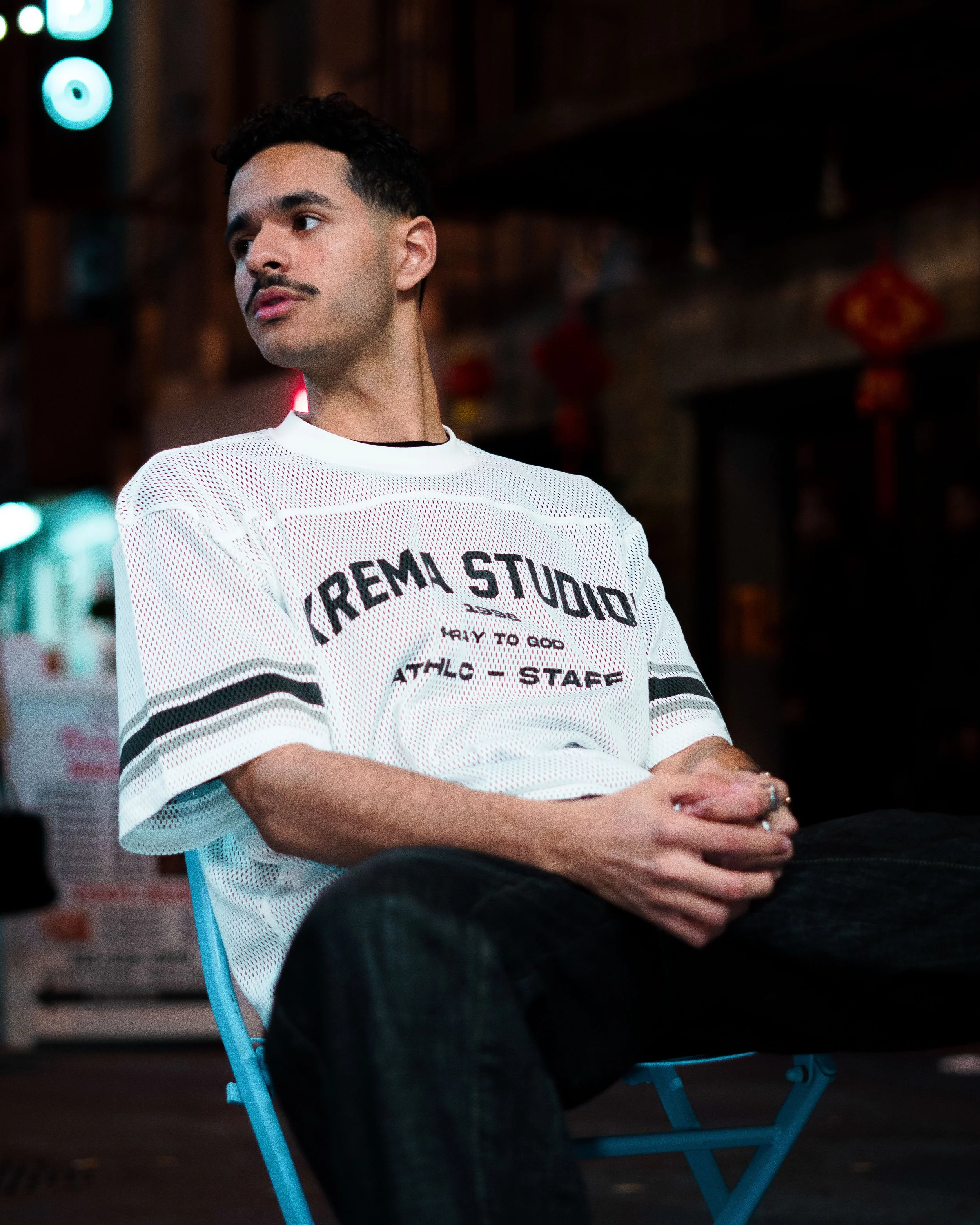 A man with dark hair and a mustache sits on a blue chair in a dimly lit space, wearing a white mesh shirt with black text.