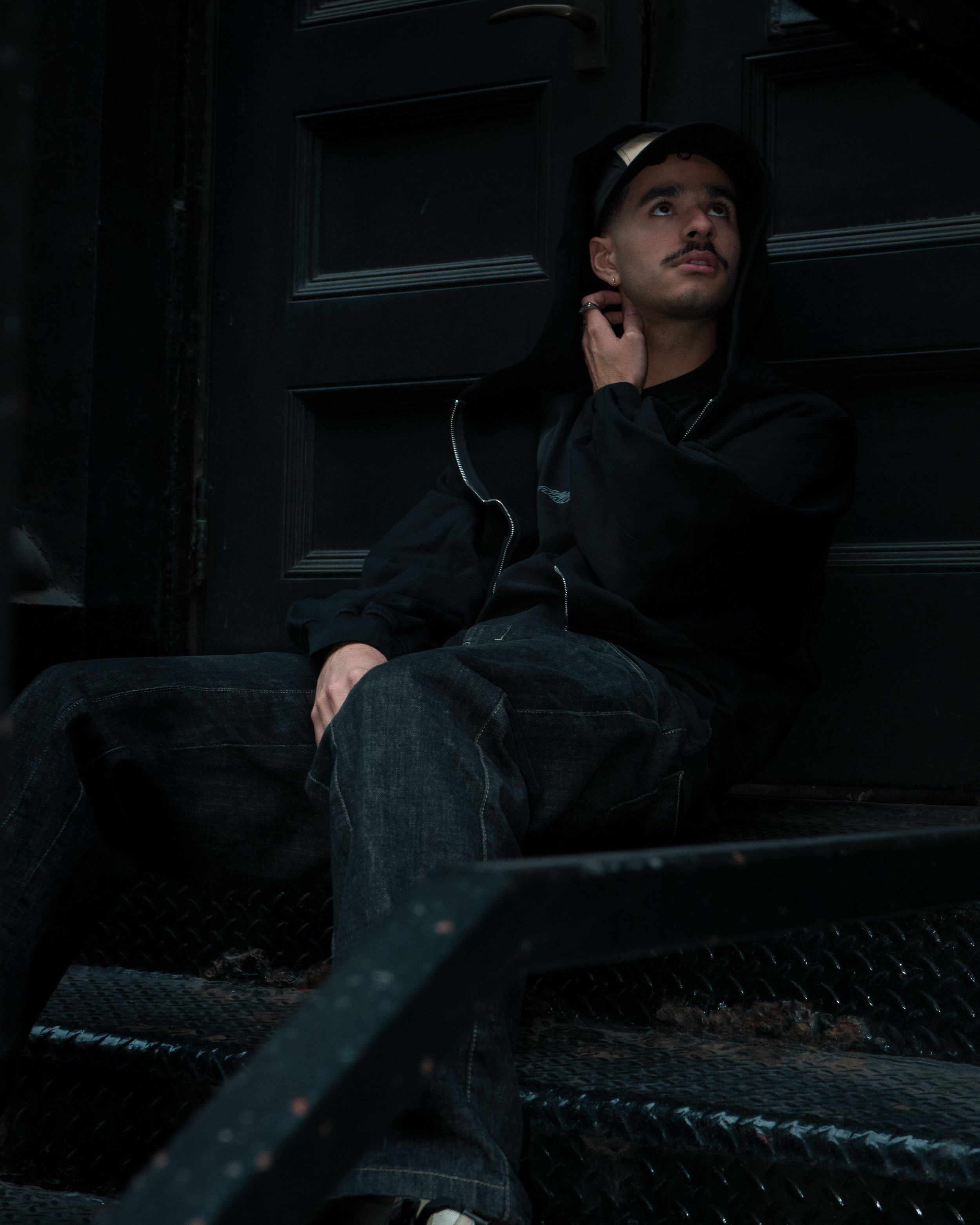 A young man in dark clothing and a helmet sits against a black door, looking upward with a contemplative expression.