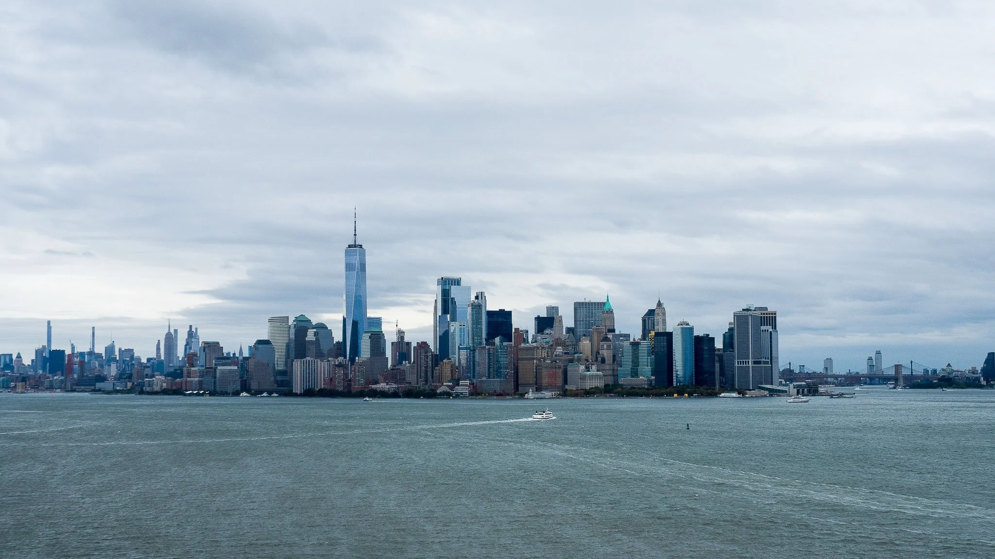 View of the New York City skyline with tall skyscrapers, including One World Trade Center, taken across a body of water with boats, under a cloudy sky.