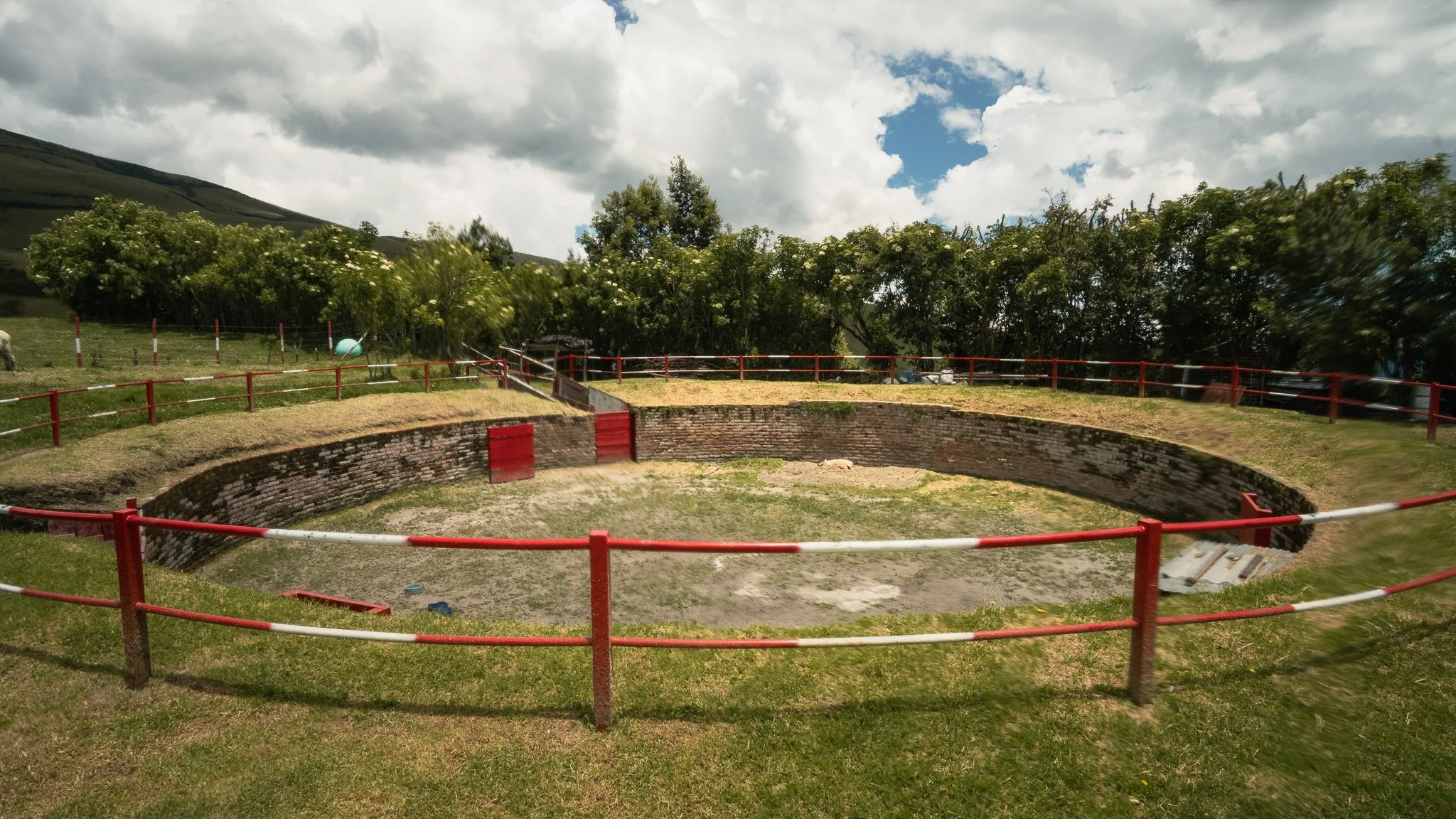 A circular structure made of brick with red and white safety barriers around it, located outdoors on grass with trees and mountains in the background under a partly cloudy sky.