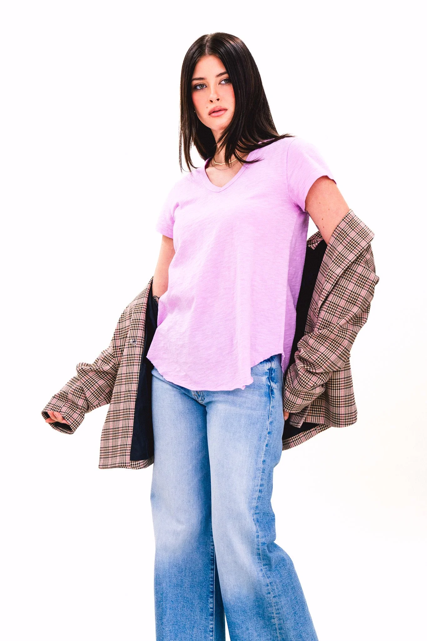 A woman with shoulder-length dark hair wearing a light purple T-shirt, ripped jeans, and a plaid blazer draped over her shoulders, standing against a white background.