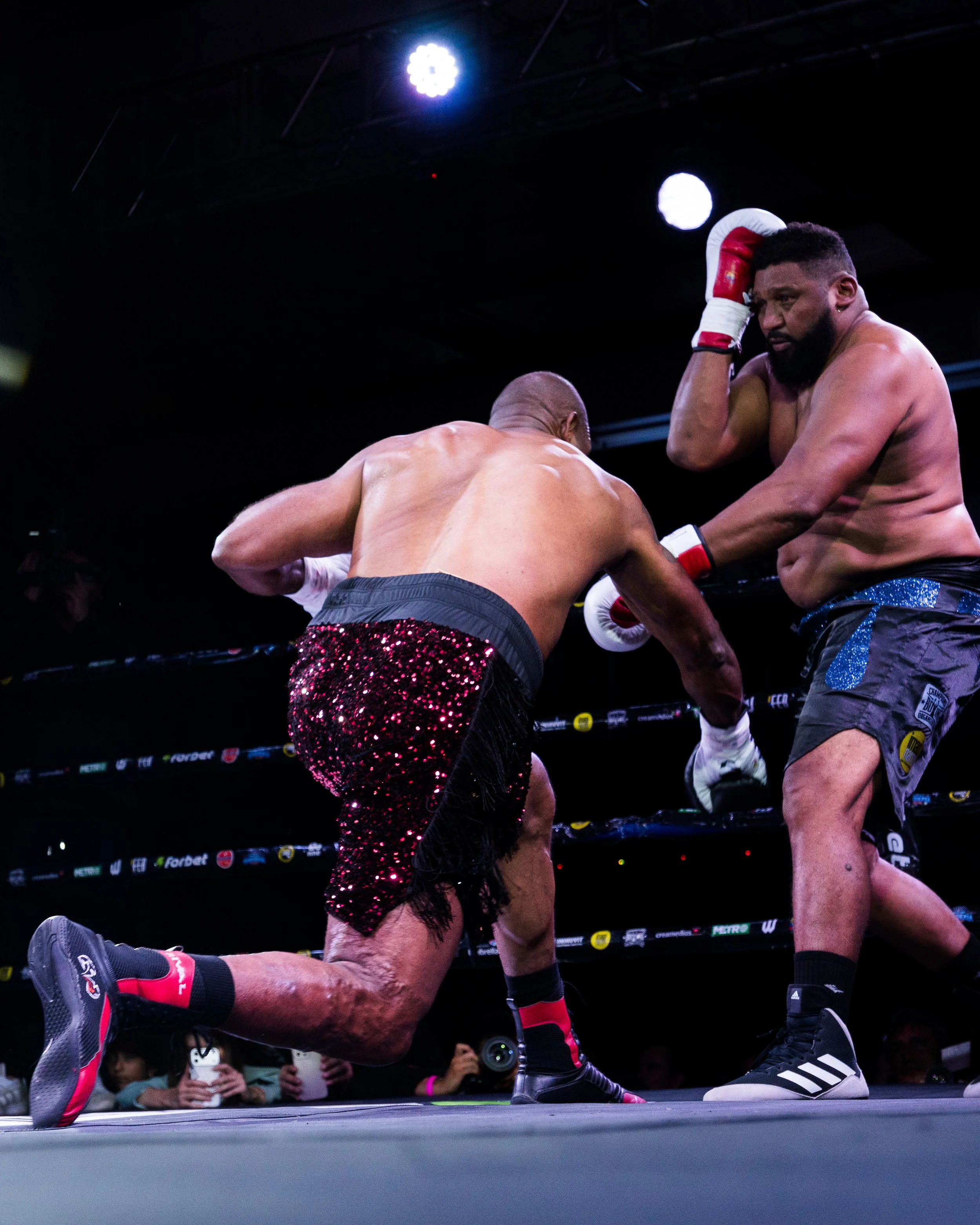 Two shirtless male boxers competing in a boxing match, with one delivering a punch to the other's body, inside a boxing ring with spectators in the background.