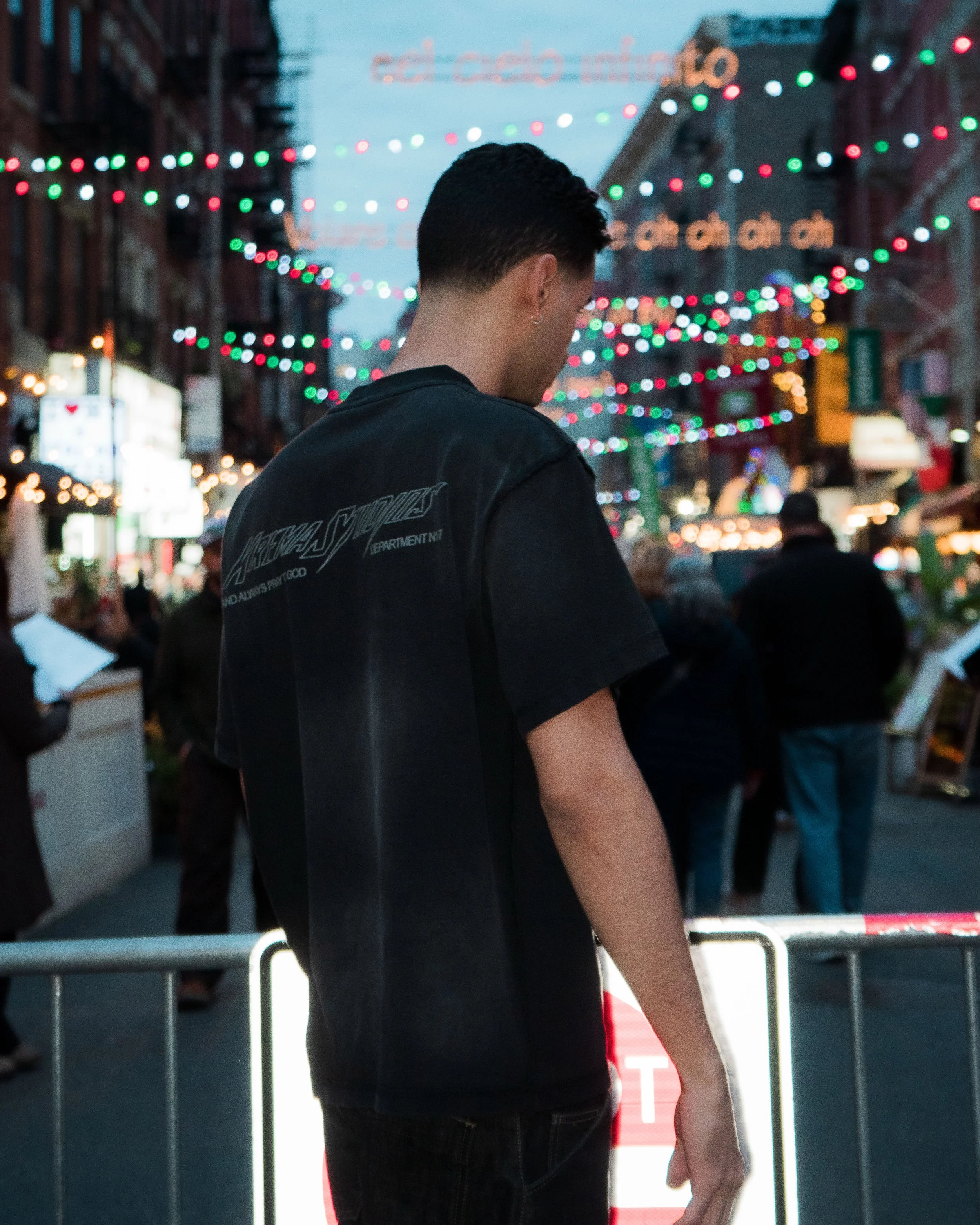 Young man with short dark hair wearing a black T-shirt standing on a city street at dusk, decorated with colorful string lights, with a crowd of people around and street signs in the background.
