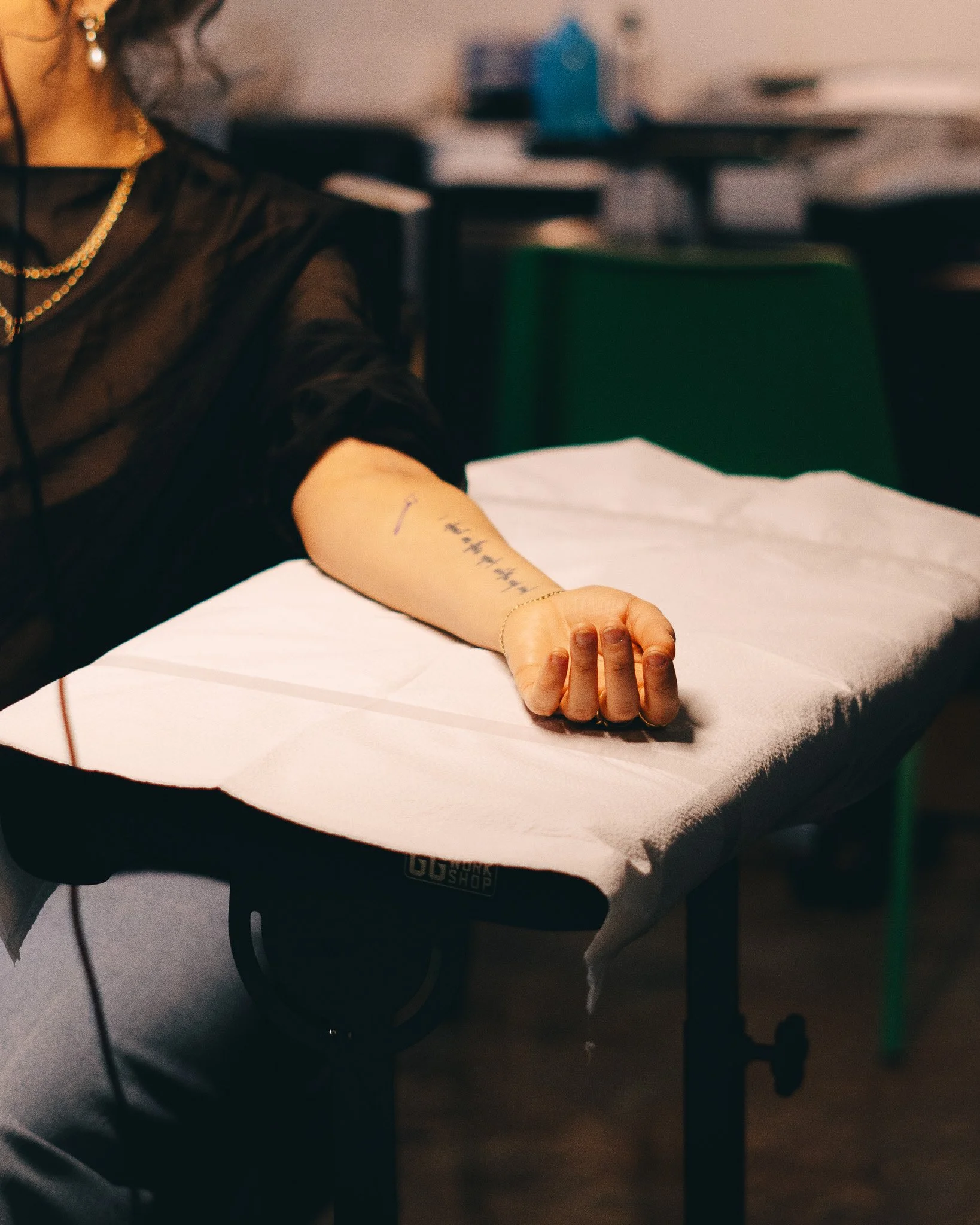 A person receiving a vaccination or injection in their arm while seated at a medical table, with paperwork and medical equipment around. The person's face is not visible, and they are wearing jewelry and dark clothing.