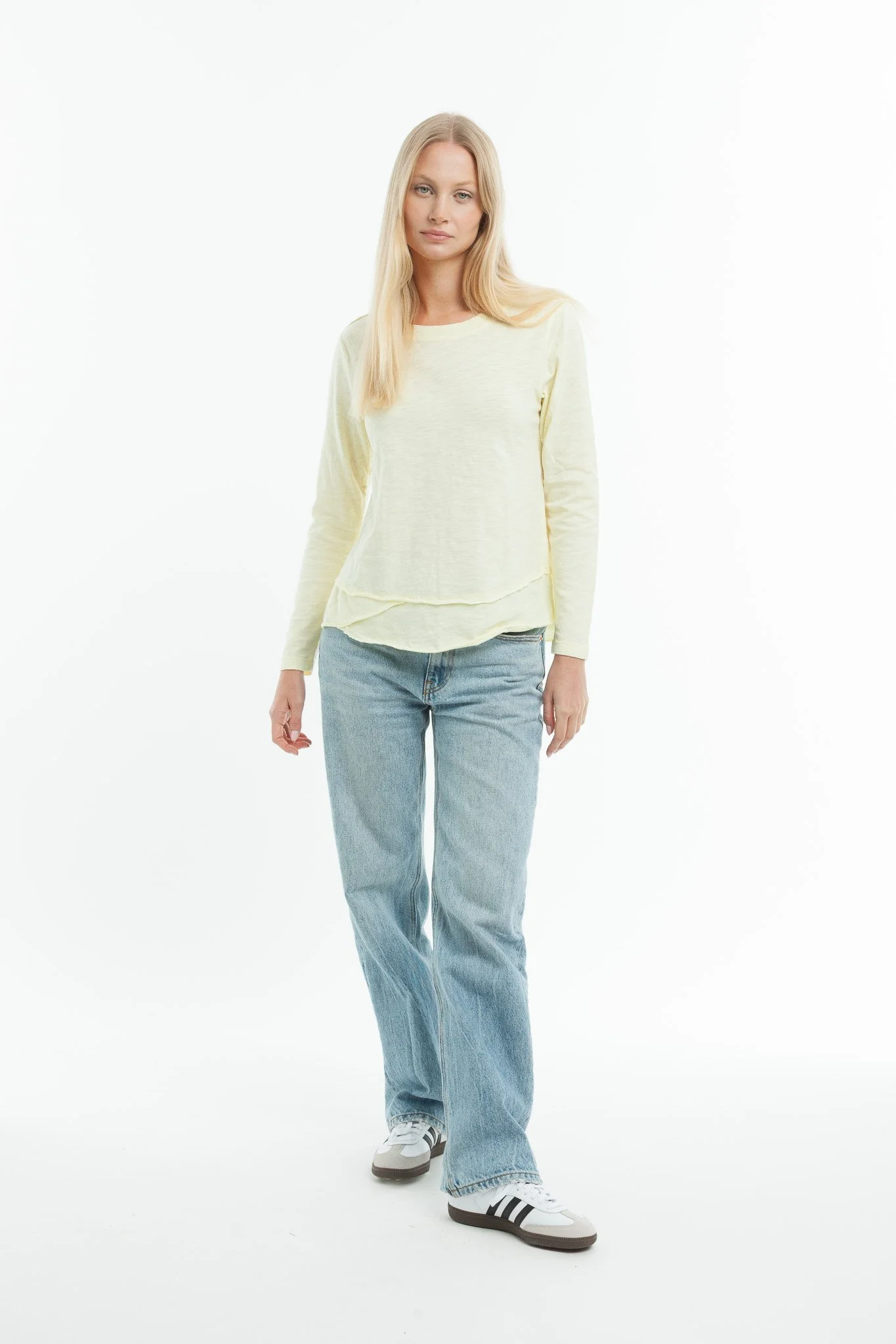 Young woman with long blonde hair in casual cream long sleeve shirt, light blue jeans, and sneakers, standing against a plain white background.
