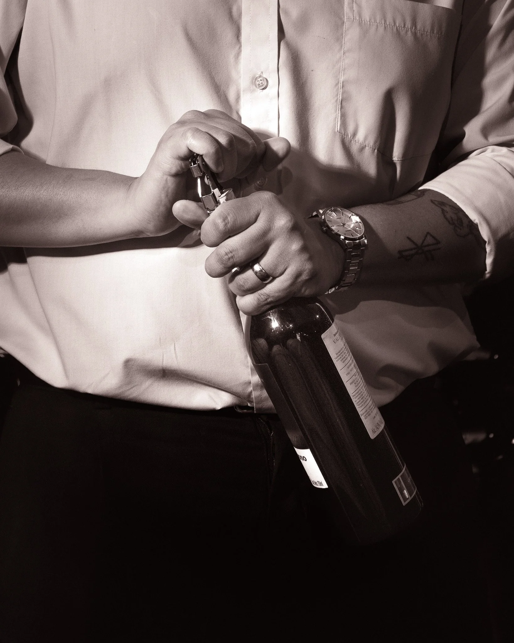 A person in a long-sleeve shirt holding a beer bottle, with a tattoo visible on their forearm, and wearing a watch. The person is clasping the bottle with both hands.
