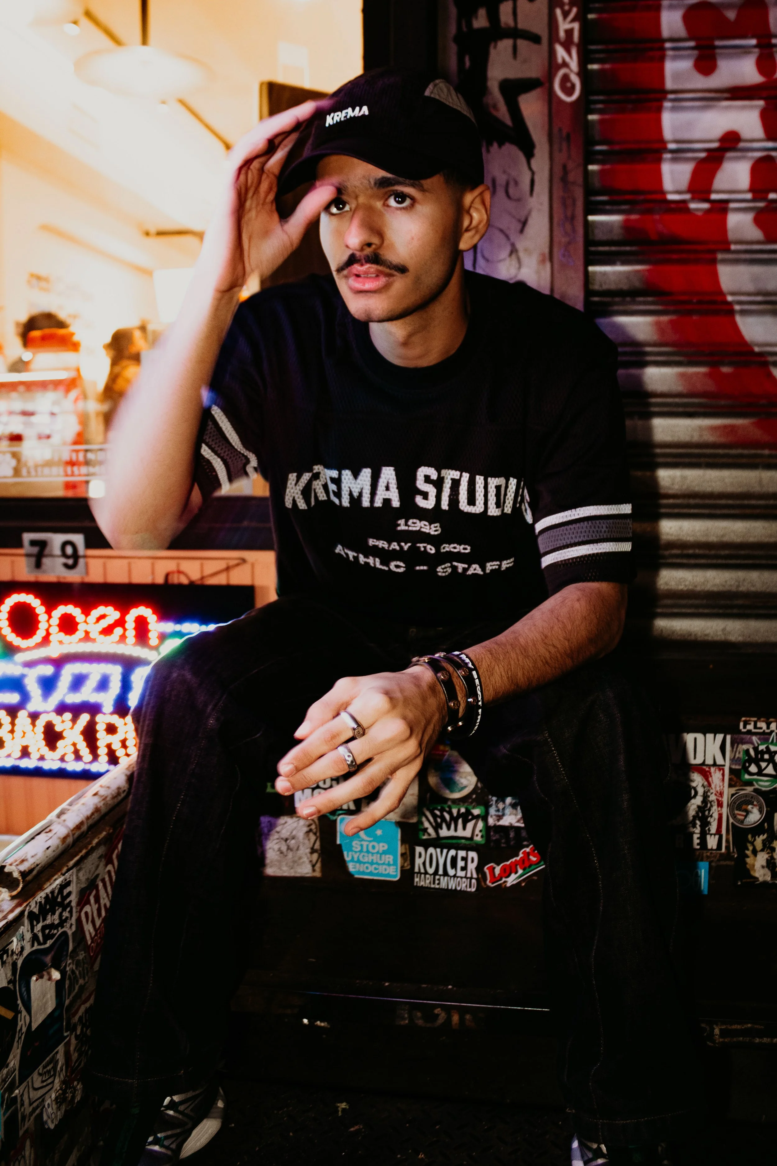 A young man is sitting on a graffiti-covered ledge at night, wearing a black t-shirt with 'KREMA STUD' printed on it and a black baseball cap, adjusting the cap with one hand. The background features red graffiti and a neon sign, with stickers on the