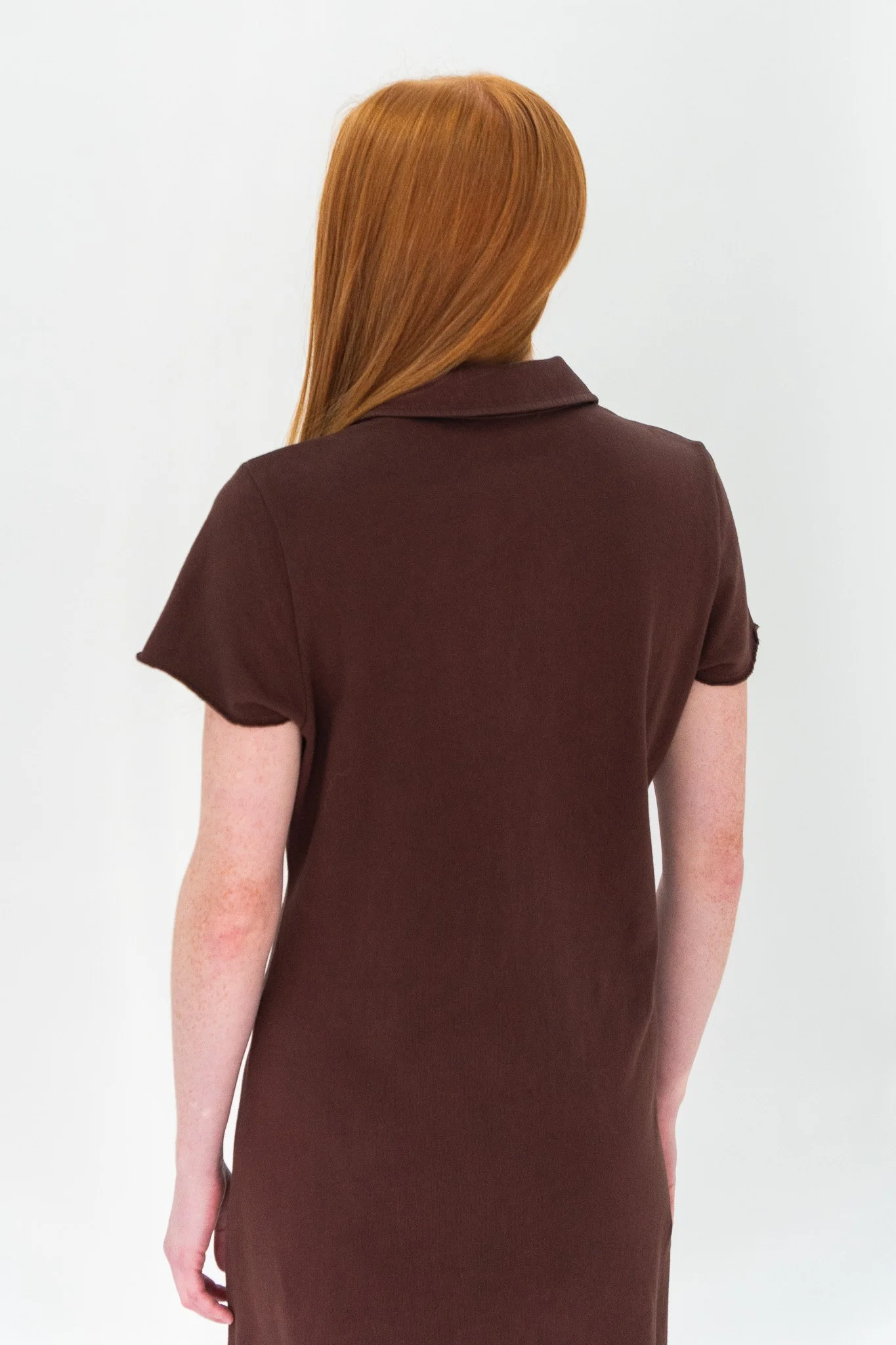 Back view of a woman with long red hair, wearing a dark brown shirt, standing against a plain white background.