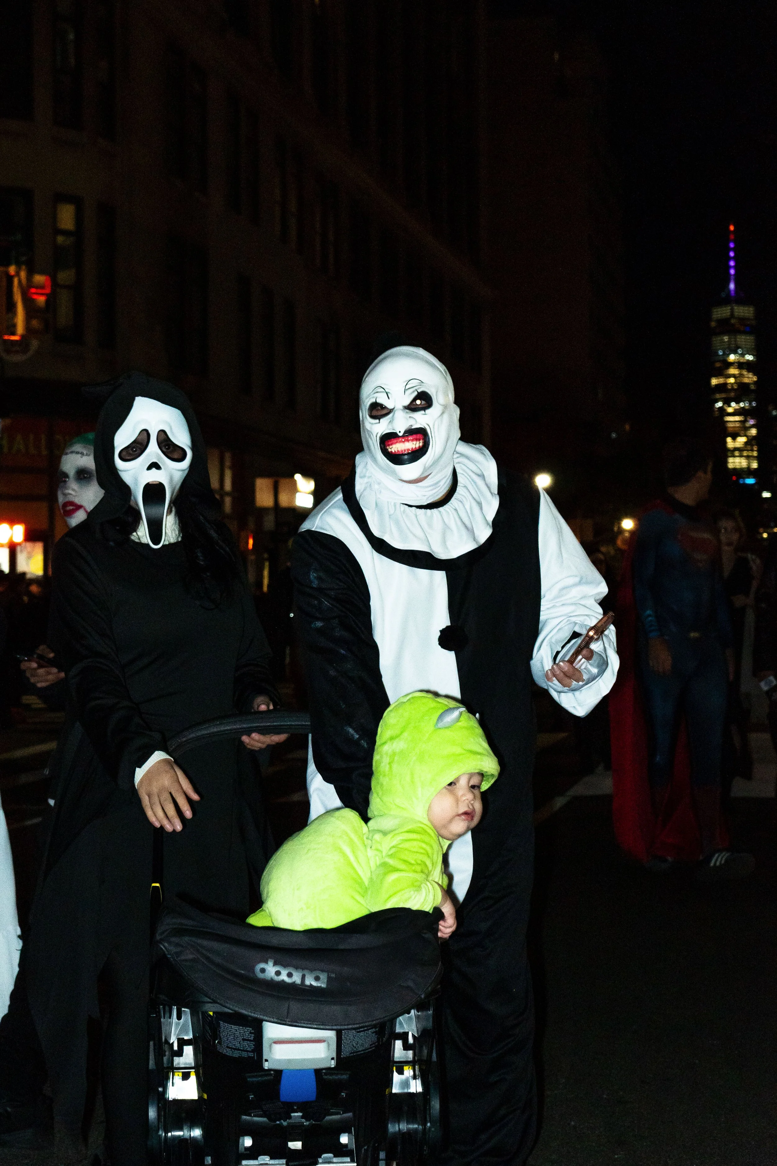 People dressed in Halloween costumes, including a person in a ghost face mask, a clown mask, and a child in a green dinosaur costume, walking on a city street at night.