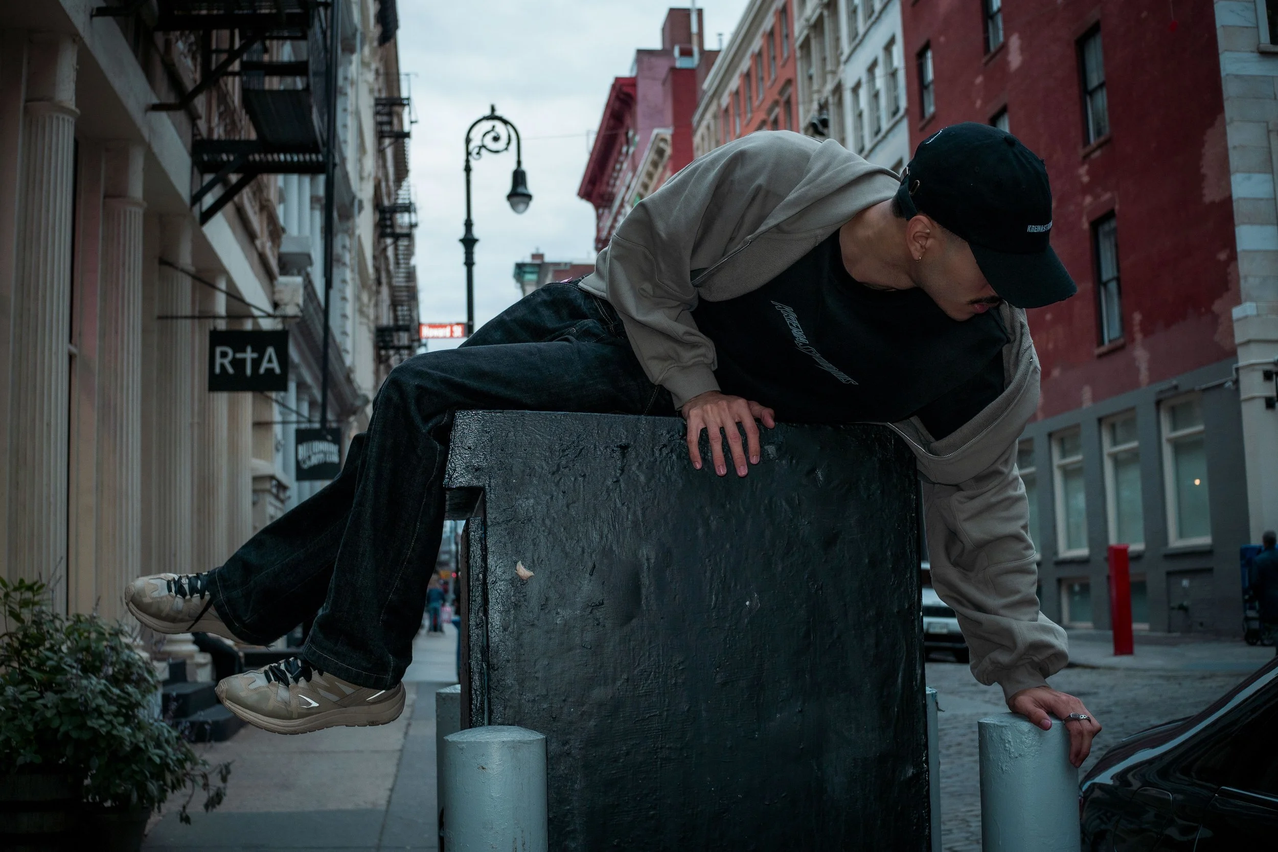 Person climbing over a city street barrier while wearing a black cap, black hoodie, beige jacket, and sneakers, with buildings and street lamps in the background.