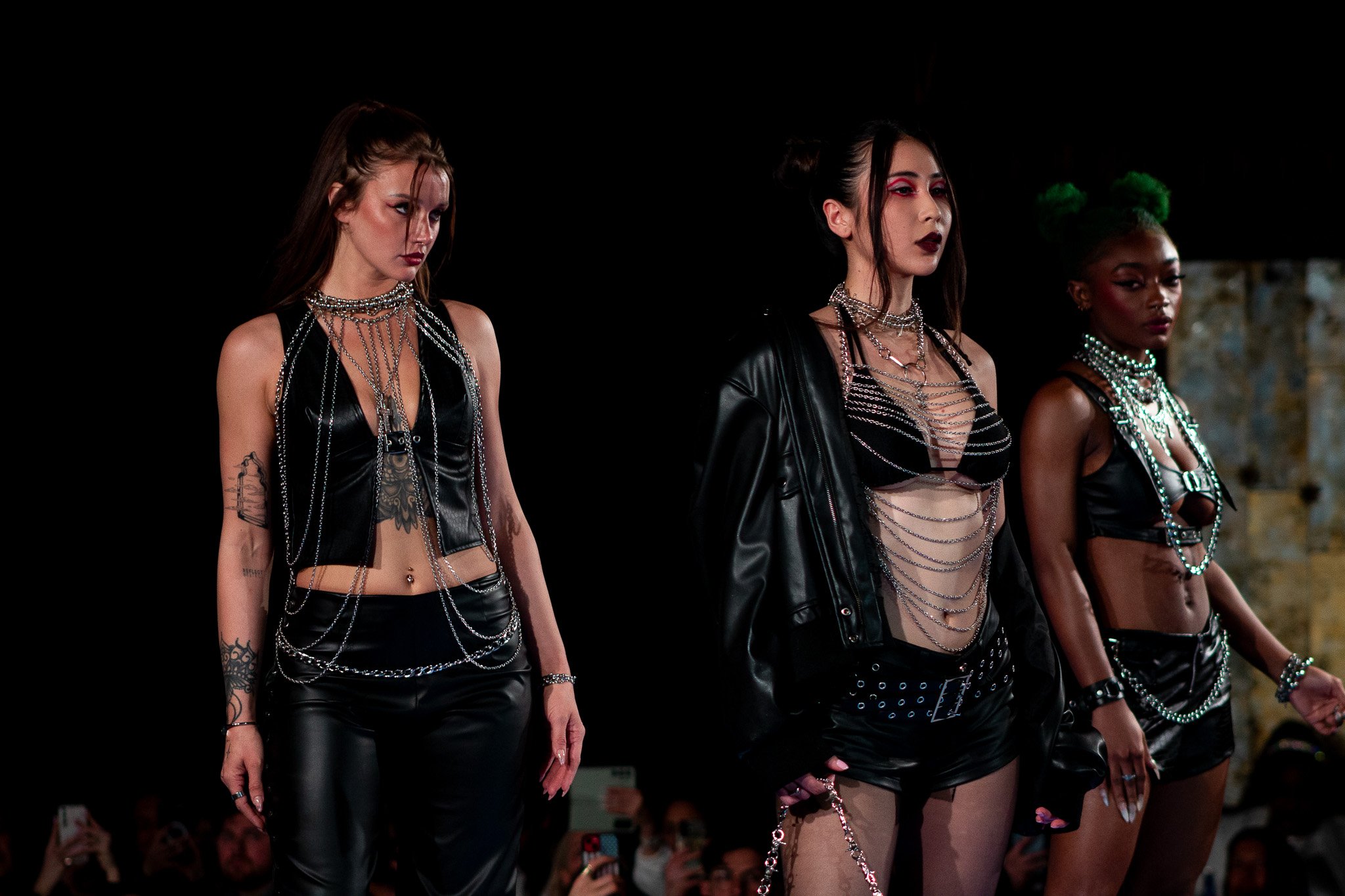 Three women standing in a row, dressed in black leather outfits and adorned with chains, at a fashion show.