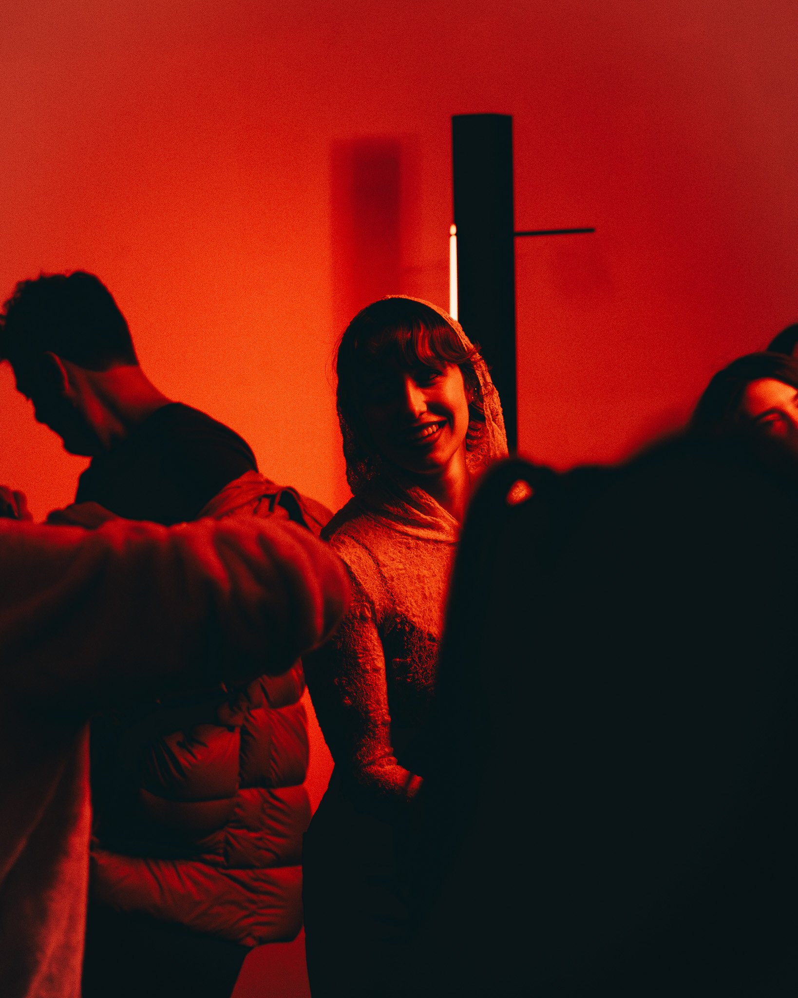 A group of people in a dark room illuminated by red lighting, with some individuals partially visible and others smiling.
