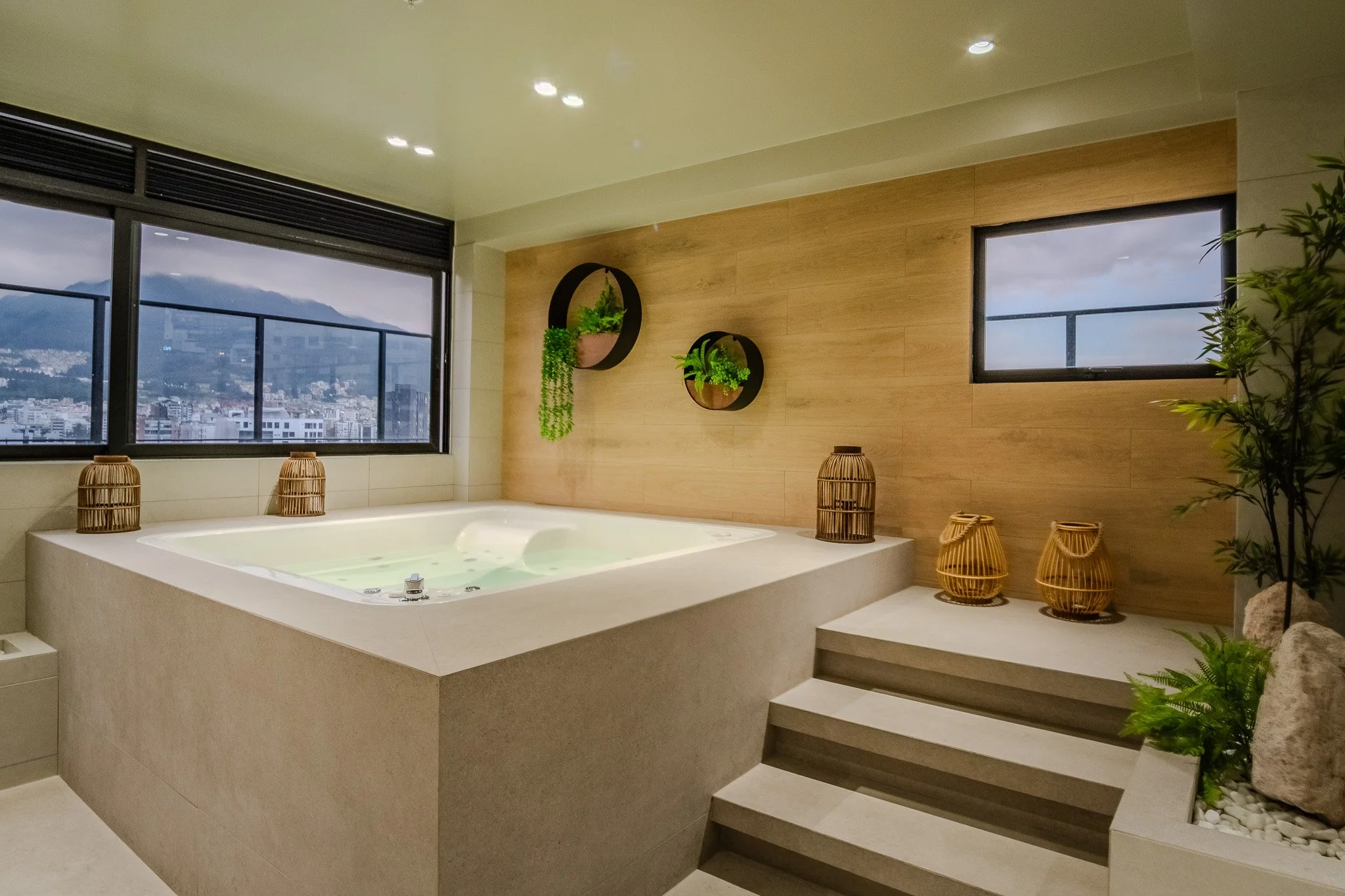Indoor hot tub surrounded by wooden and stone decor, with large windows offering city and mountain views, and decorative plants and baskets nearby.