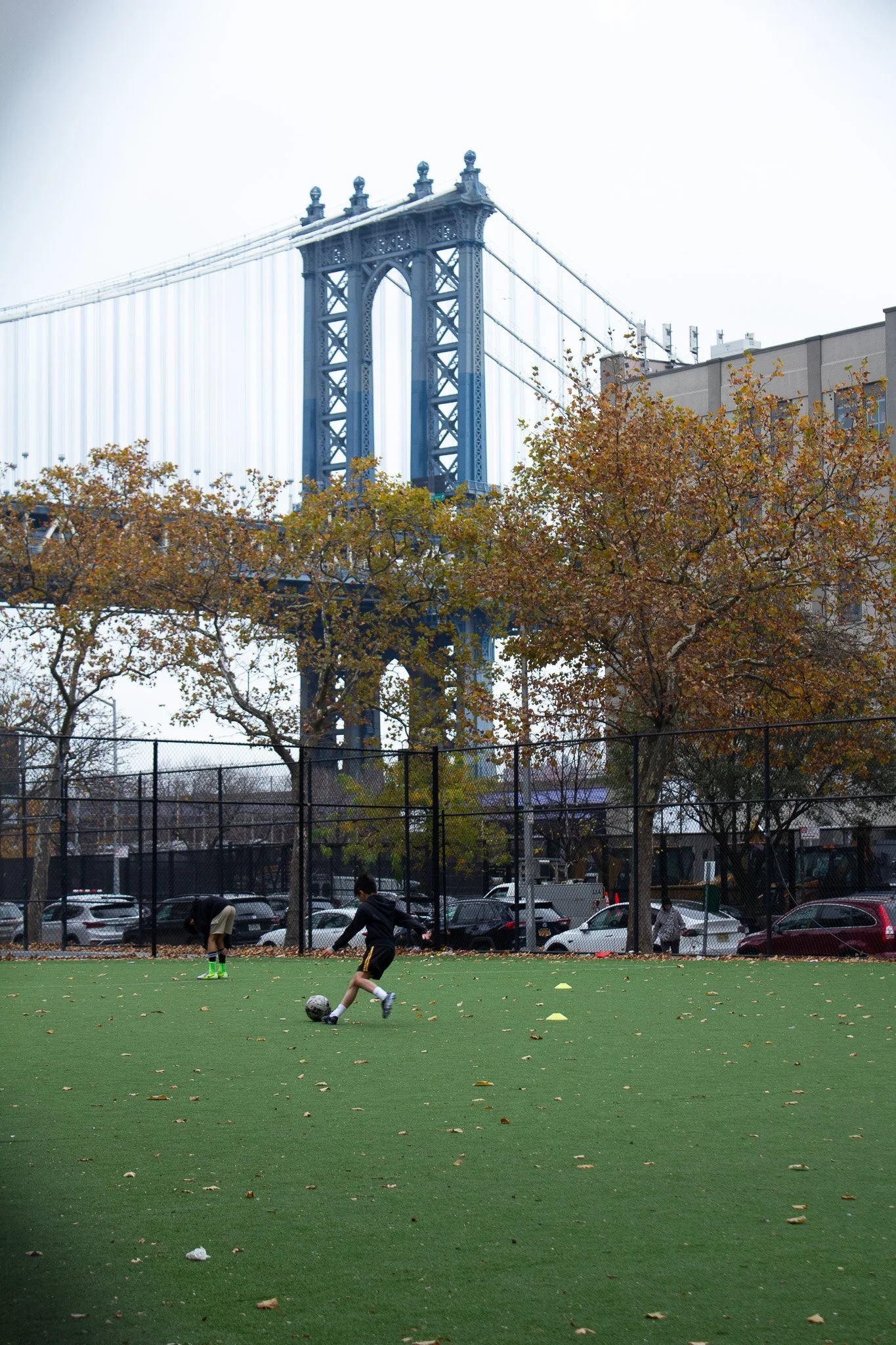 Children playing soccer on a green field with a large bridge in the background and autumn trees around.