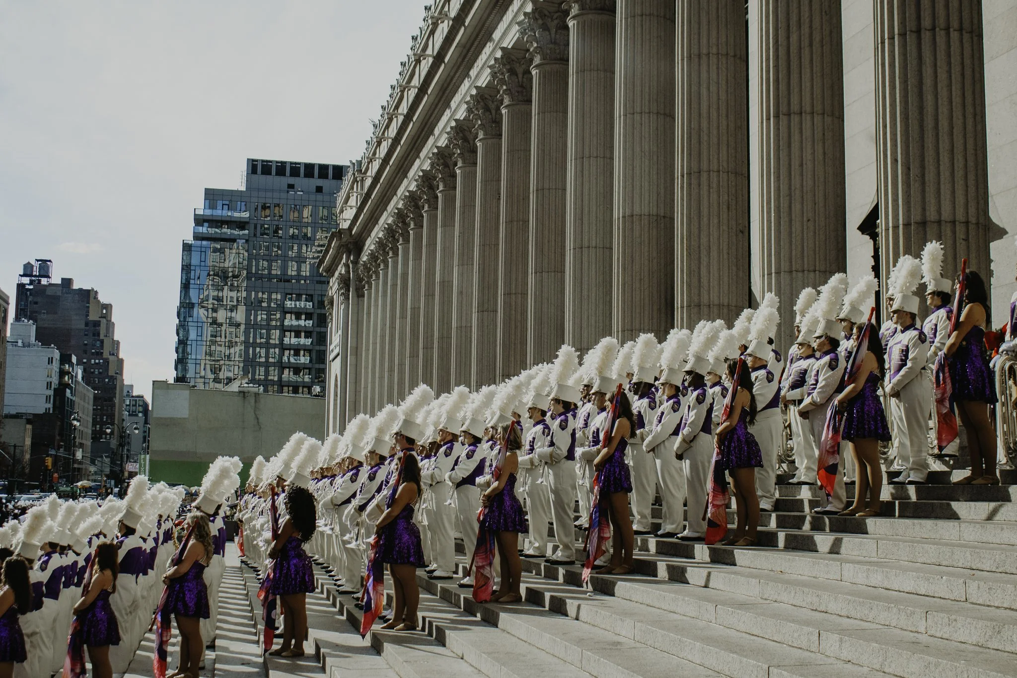 Group of uniformed individuals with white hats and purple outfits standing in formation on the steps of a grand building with large pillars, city skyscrapers in the background.