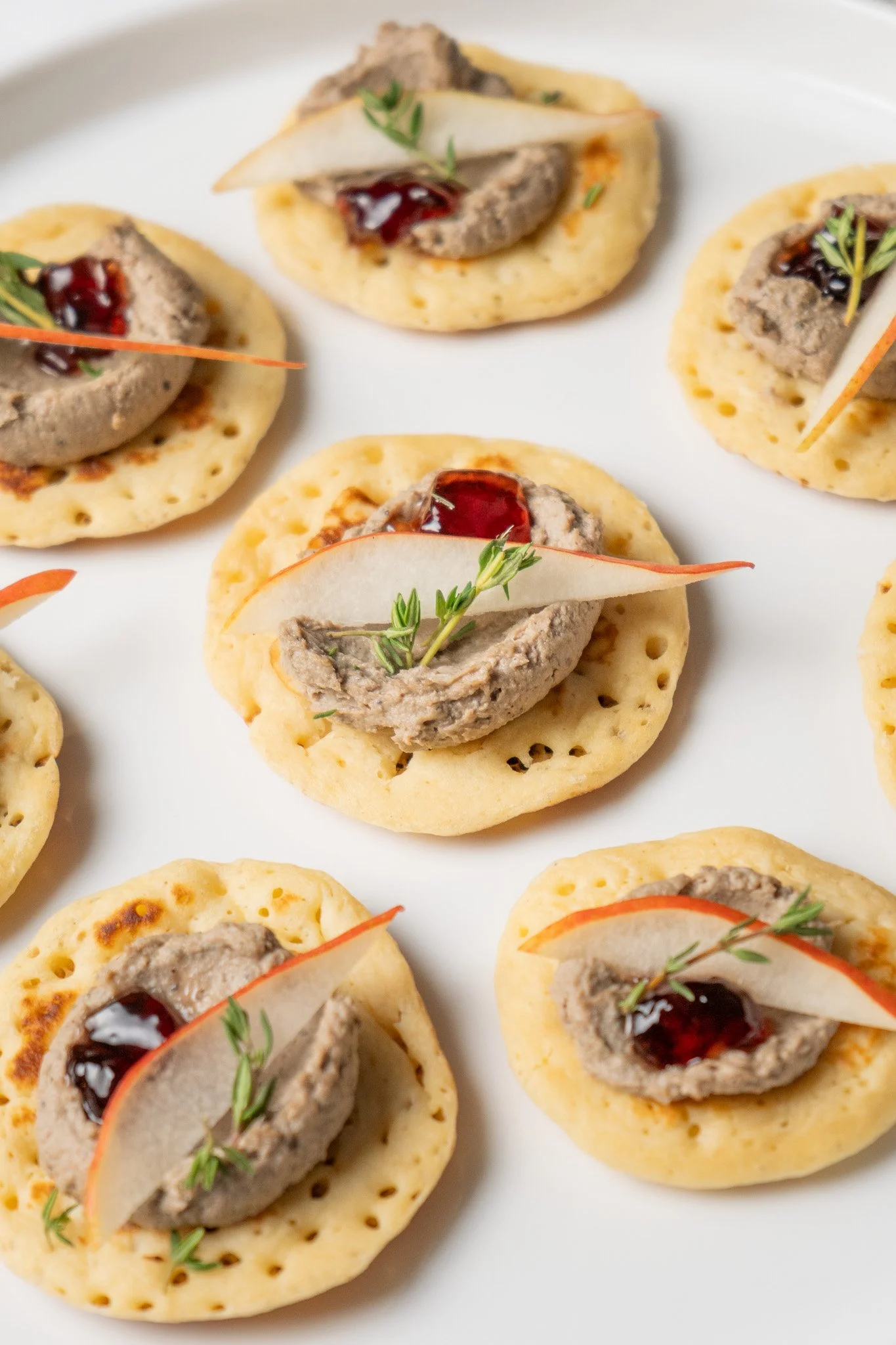 Appetizer bites with a cracker base topped with a dollop of pâté, garnished with a thin slice of pear, a small amount of red jam, and green herbs.