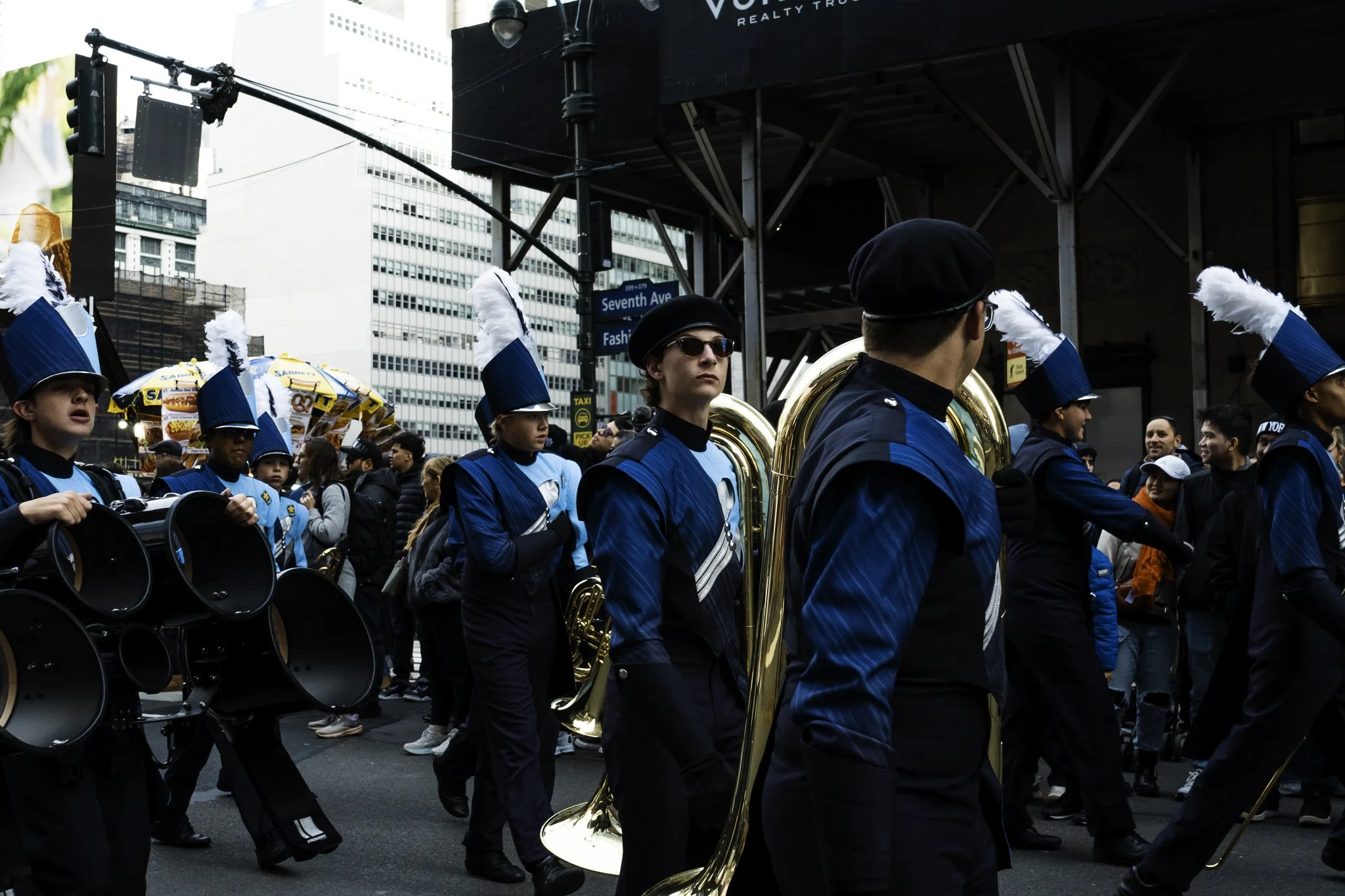 A marching band in parade uniform playing musical instruments as they march down a city street, with onlookers in the background and tall buildings around.