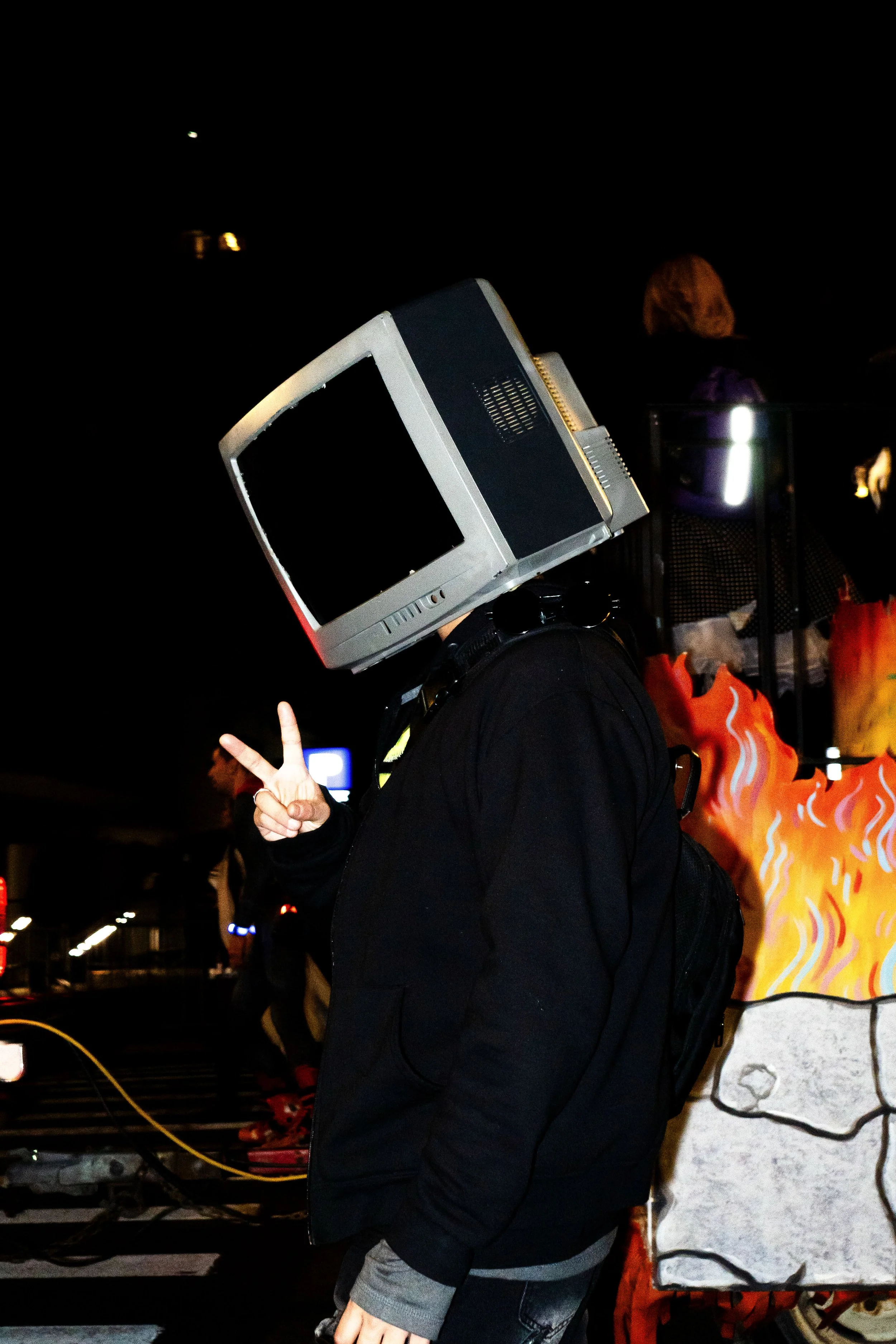 Person wearing a vintage computer monitor as a helmet making a peace sign at night.