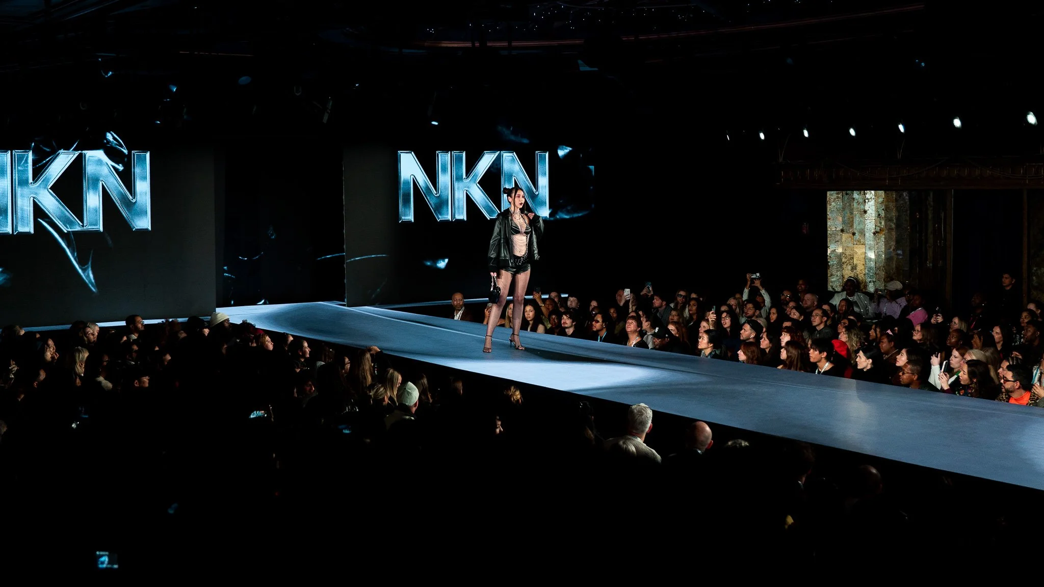 A model walking on a runway at a fashion show, wearing a black outfit with heels and holding a small purse, while an audience watches. Large screens display the letters 'NKN' in the background.