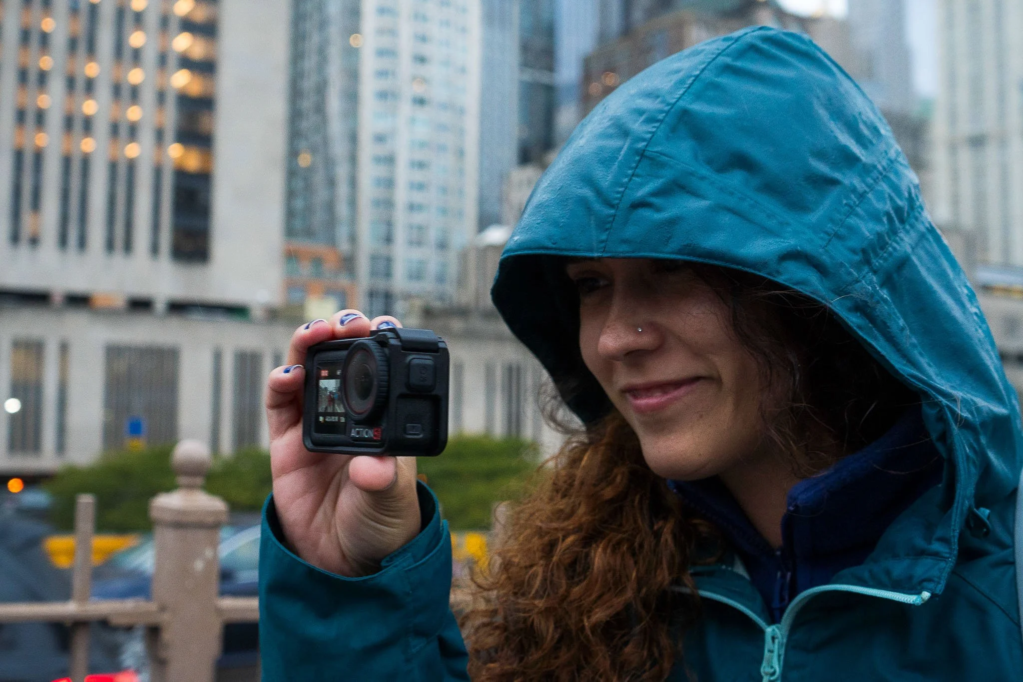 Woman in a blue hooded jacket taking a photo with a camera outdoors in the city with tall buildings in the background.