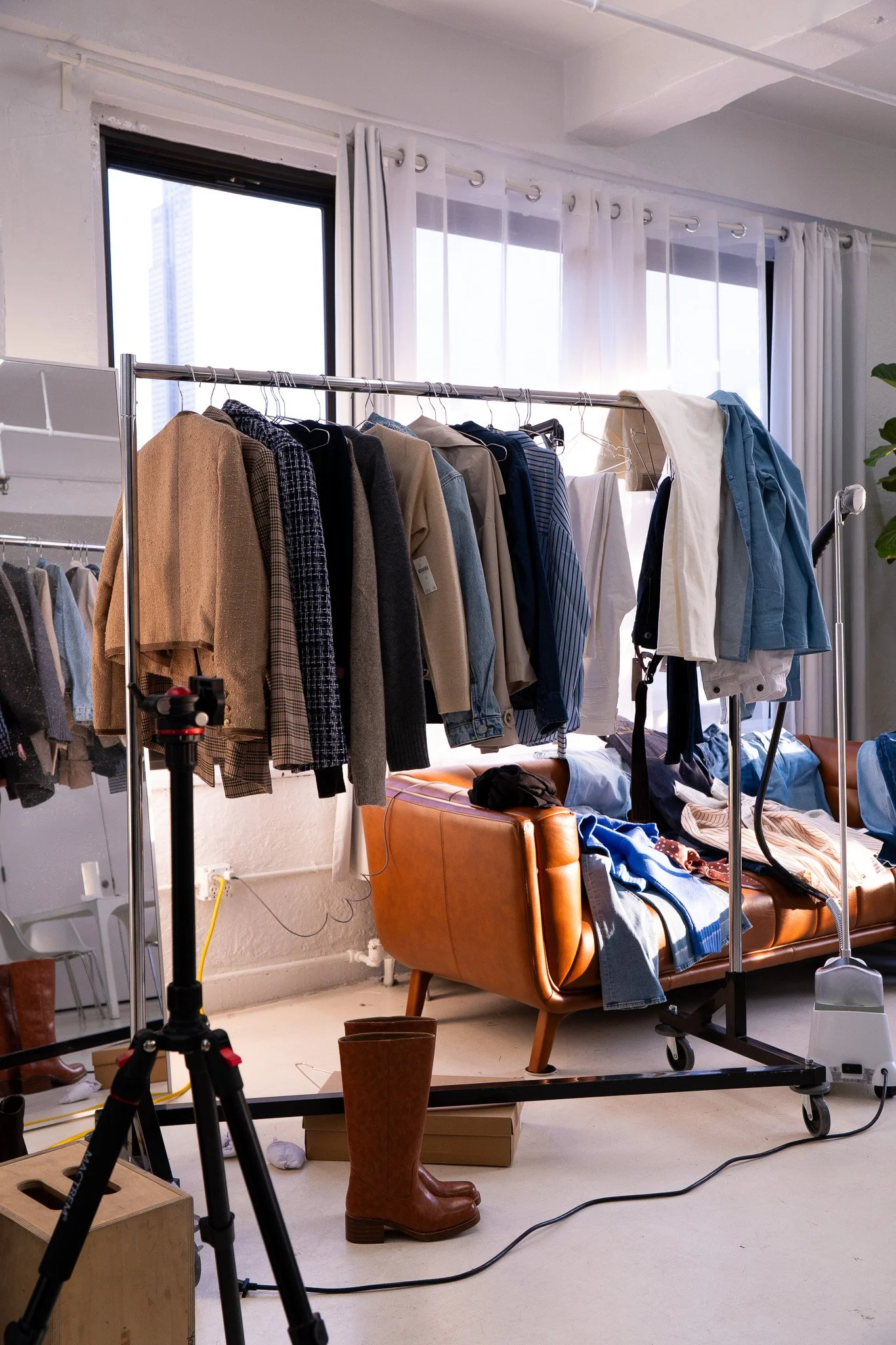 A clothing rack with various jackets and shirts in front of large windows with white curtains. A leather sofa with clothes and a pair of brown boots underneath, and a tripod with camera equipment in the foreground.