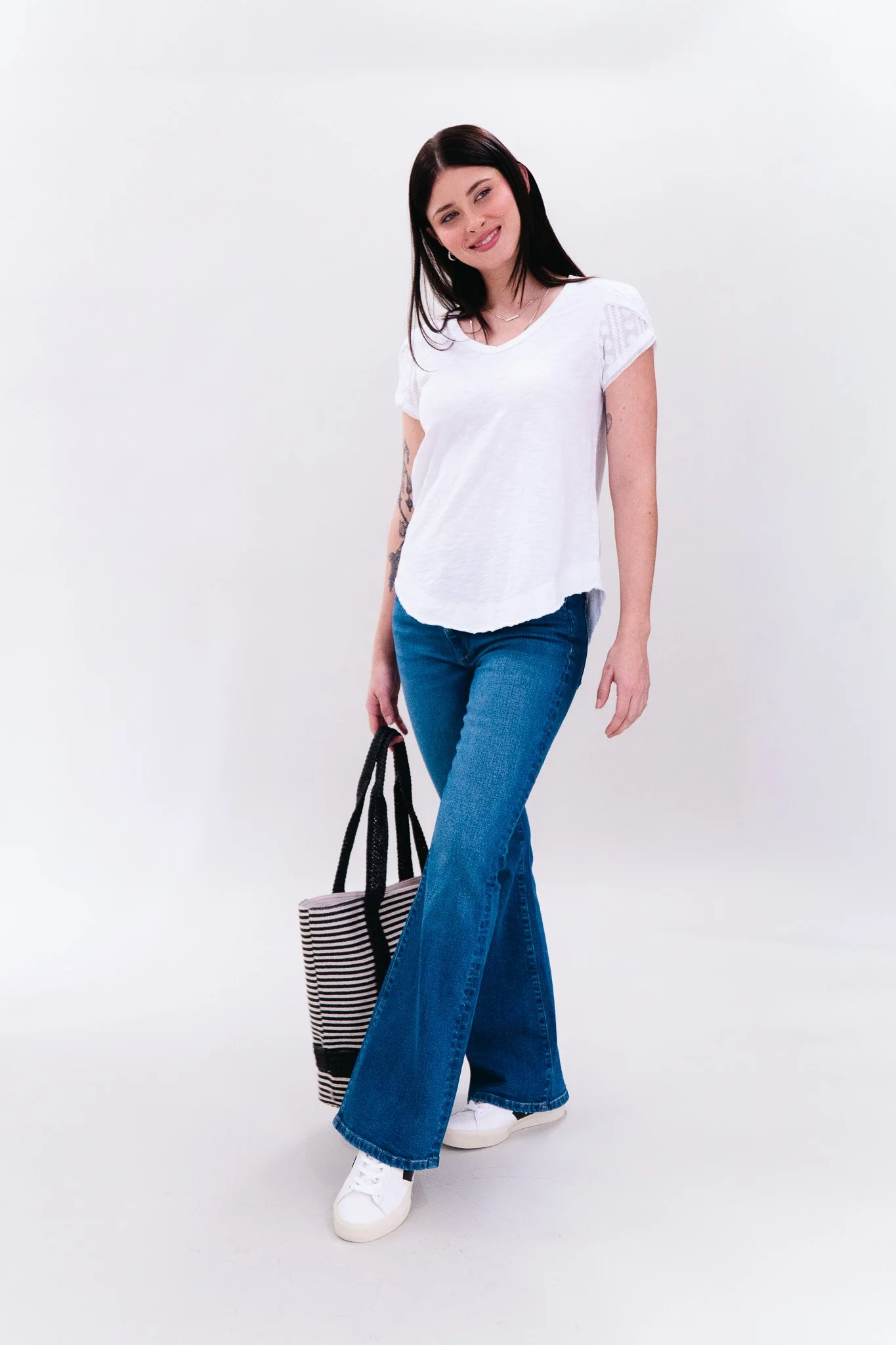 A woman stands against a white background, wearing a white t-shirt, wide-leg blue jeans, white sneakers, and holding a striped tote bag. She has dark hair and is smiling.