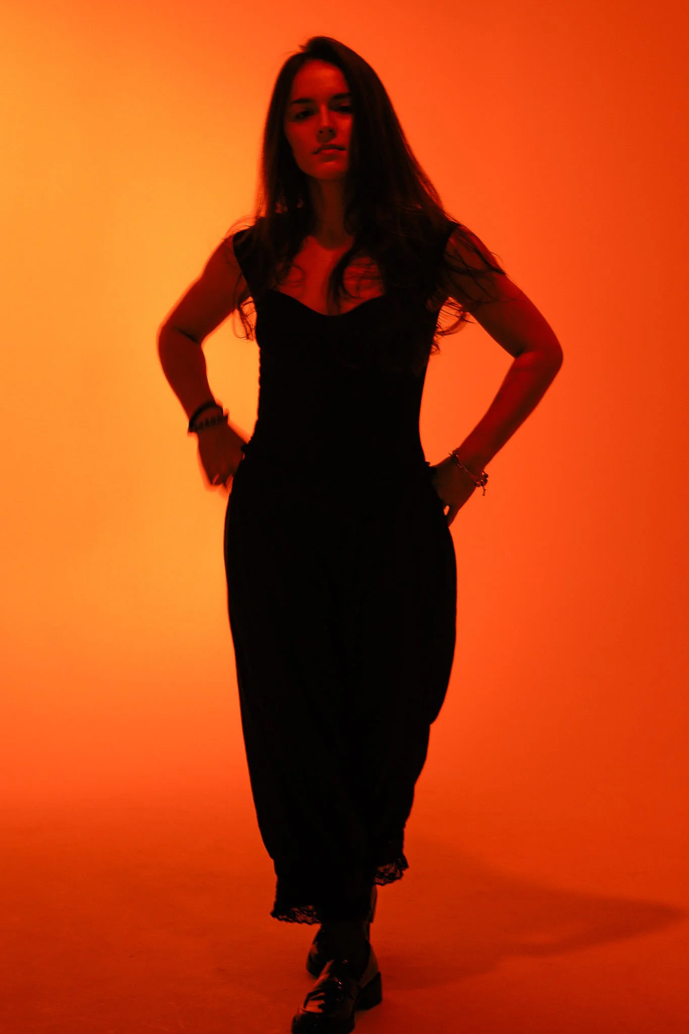 A woman with long dark hair wearing a black dress and boots standing with her hands on her hips against an orange background.